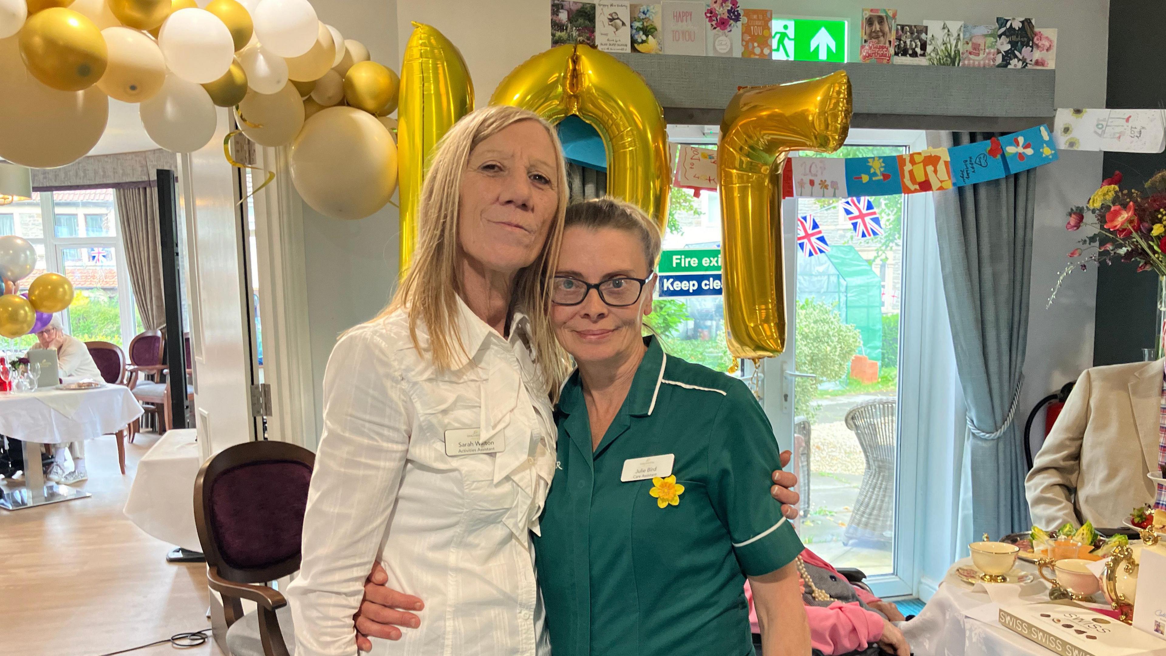 Sarah wearing a white shirt and standing beside Julie who is wearing green scrubs, glasses and a daffodil pin. Behind them there are lots of balloons and a large 107 balloon