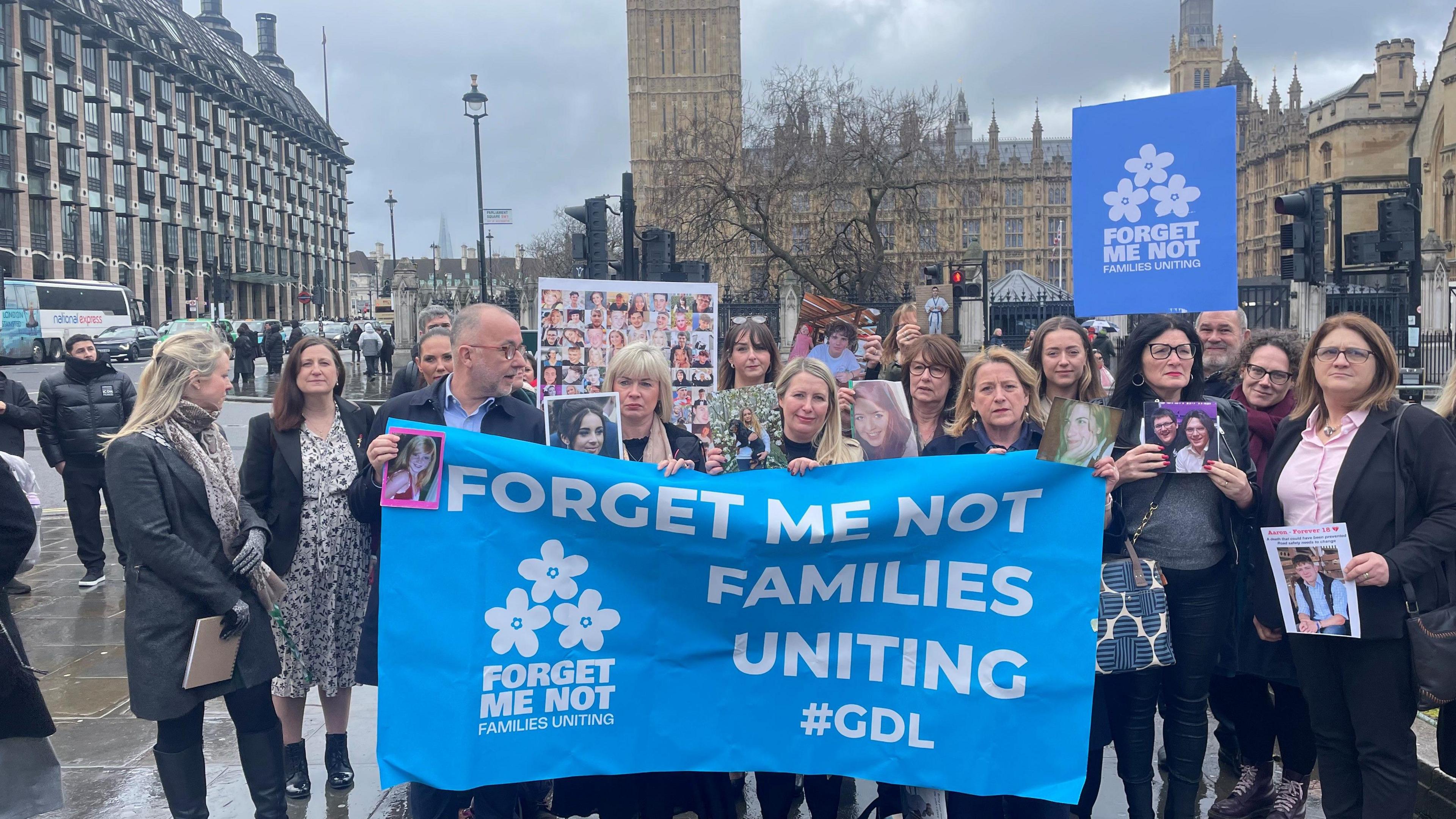 Around 20 men and women pose outside the House of Commons holding a large blue banner displaying their group's name, 'Forget Me Not Families Uniting' in large white lettering. The skies are grey and nearby traffic can be seen in the background