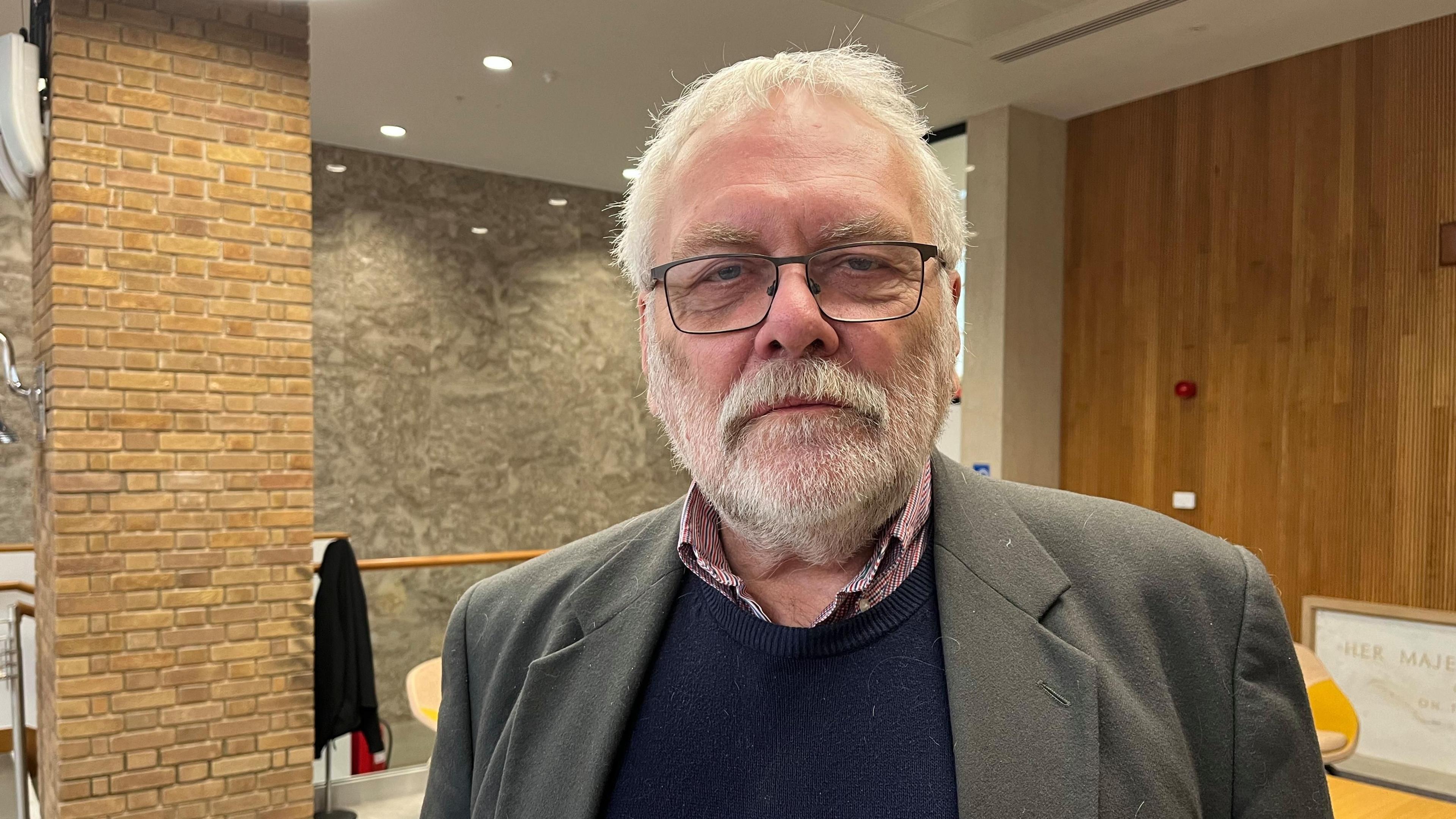 Steve Morphew has short, white hair and a white beard. He is wearing glasses and looking at the camera with a council chamber behind him.
He is wearing a jacket with a blue jumper underneath and a red checked collared shirt underneath.