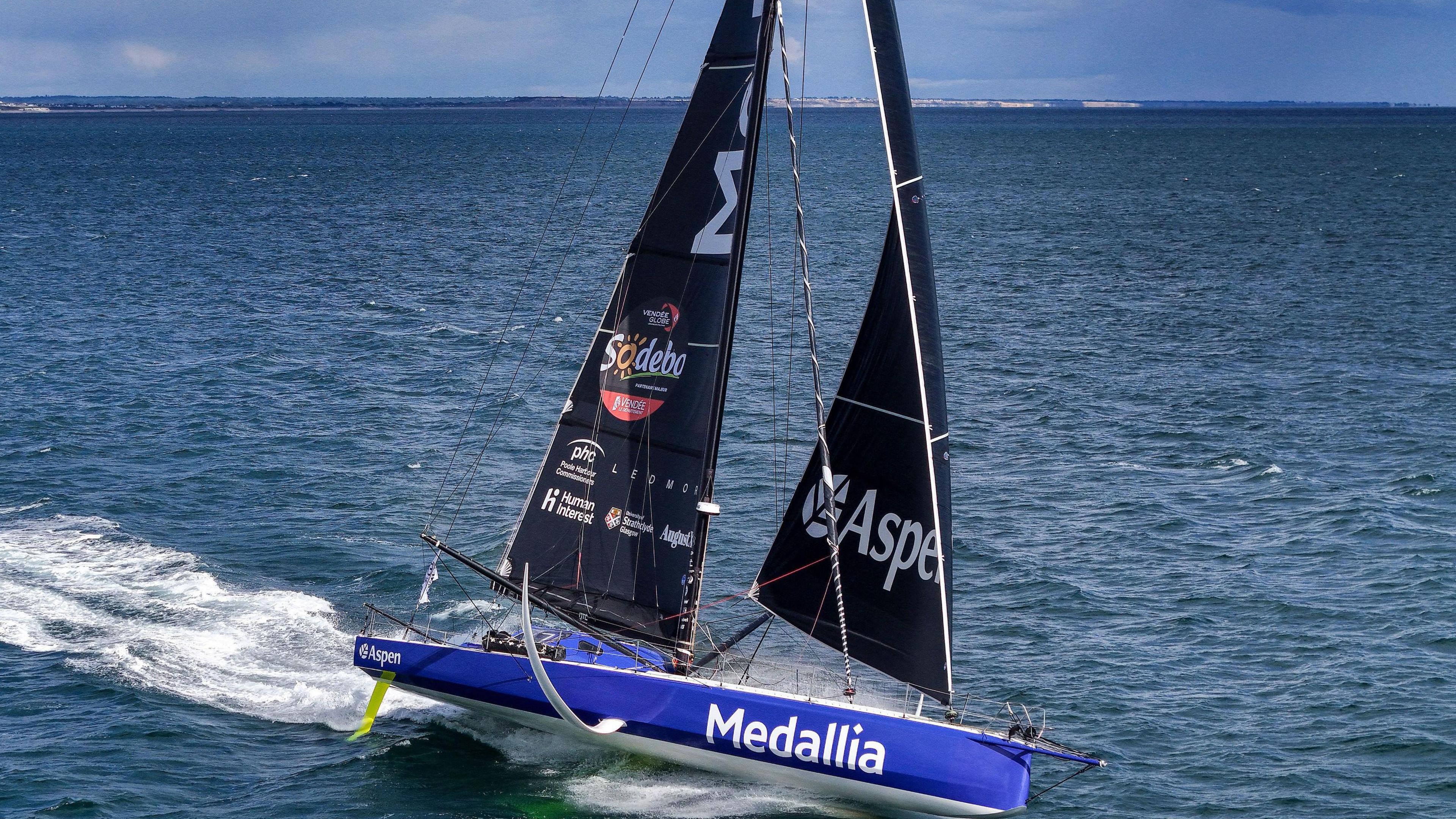 A drone image of the Medallia yacht sailing on the sea. It has large black sails and a blue body. The name Medallia is printed on the side of the yacht's body in white lettering. 