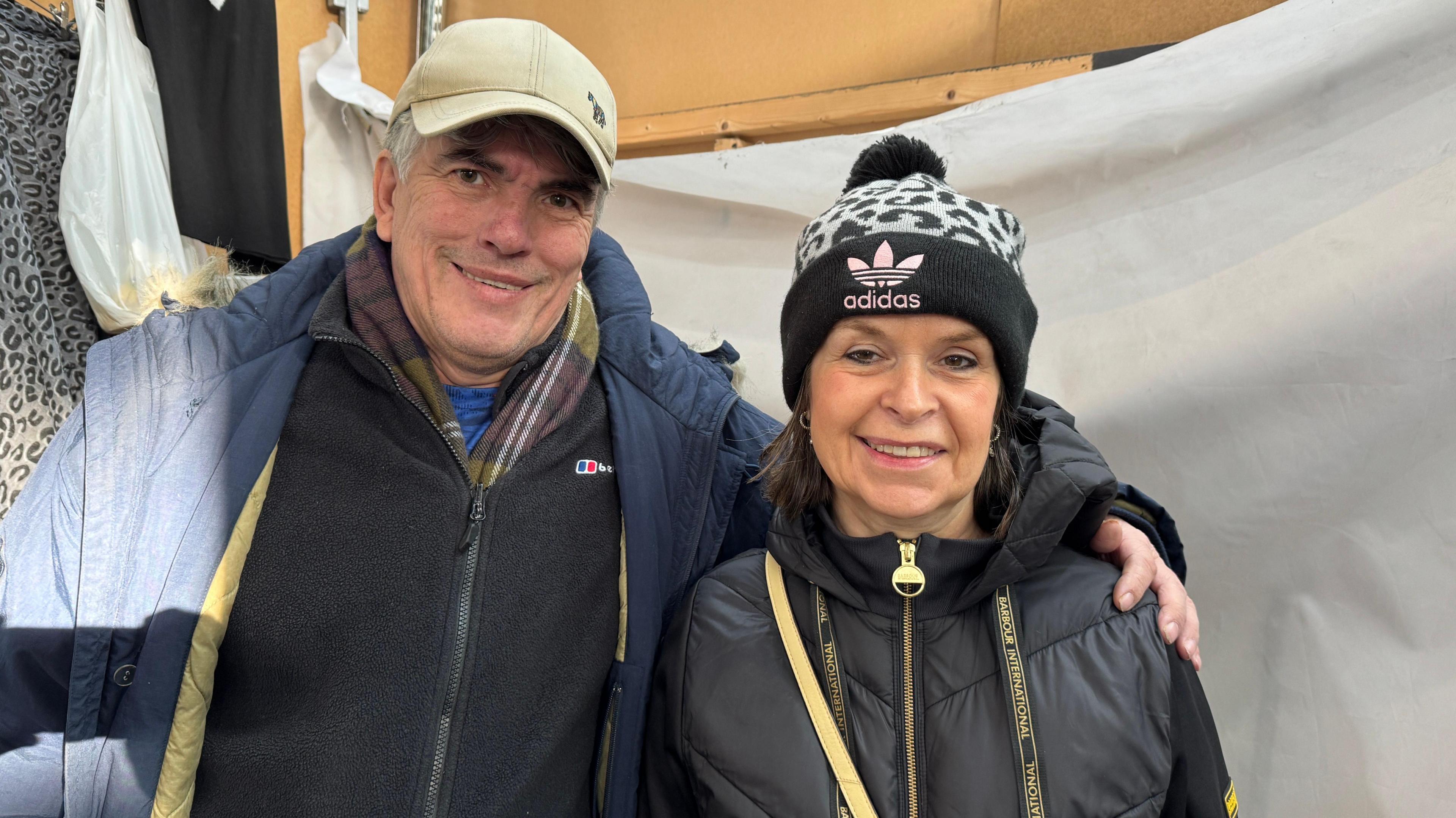 The couple are stood together with Andrew's arm around Julie. Both have dark coloured, thick winter coats on and Andrew has a brown checked scarf tucked into a fleece under his coat. Andrew has a beige cap on and Julie a black patterned bobble hat.