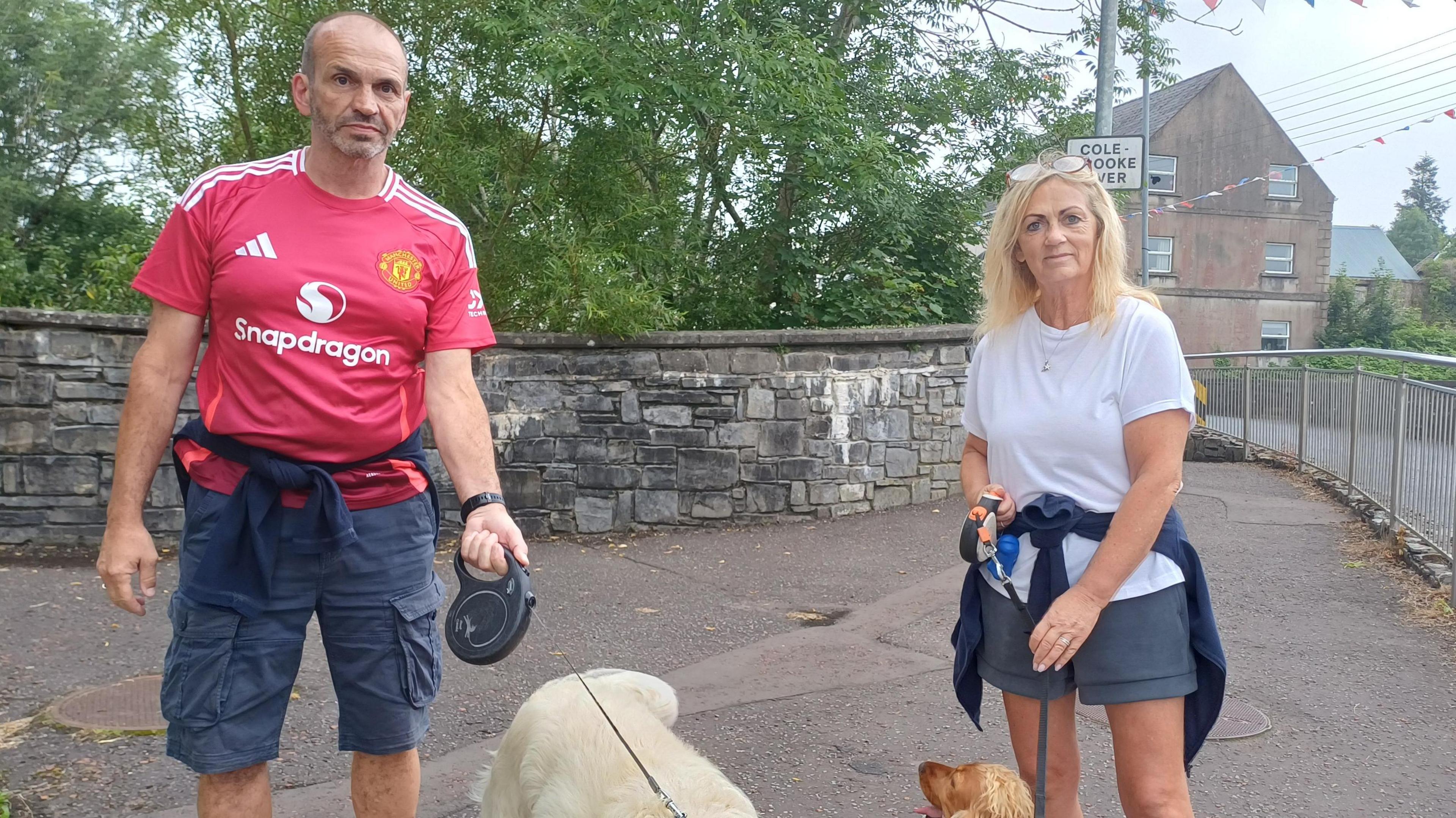 Two people holding dog leads. A man on the left is wearing a red Man United football shirt. A woman, right, is wearing a white t-shirt and has long blonde hair.