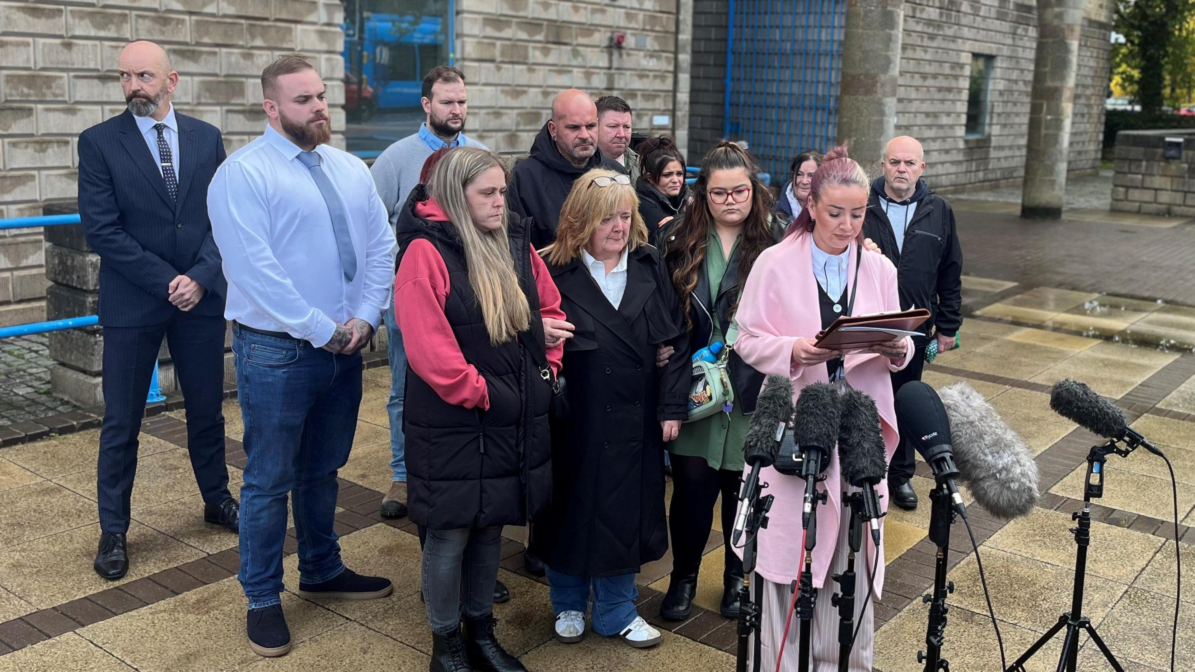 A group of people gathered outside a court building with sombre expressions on their faces. One woman wearing a long pink coat is reading from a document in her hands, while standing in front of multiple large microphones.