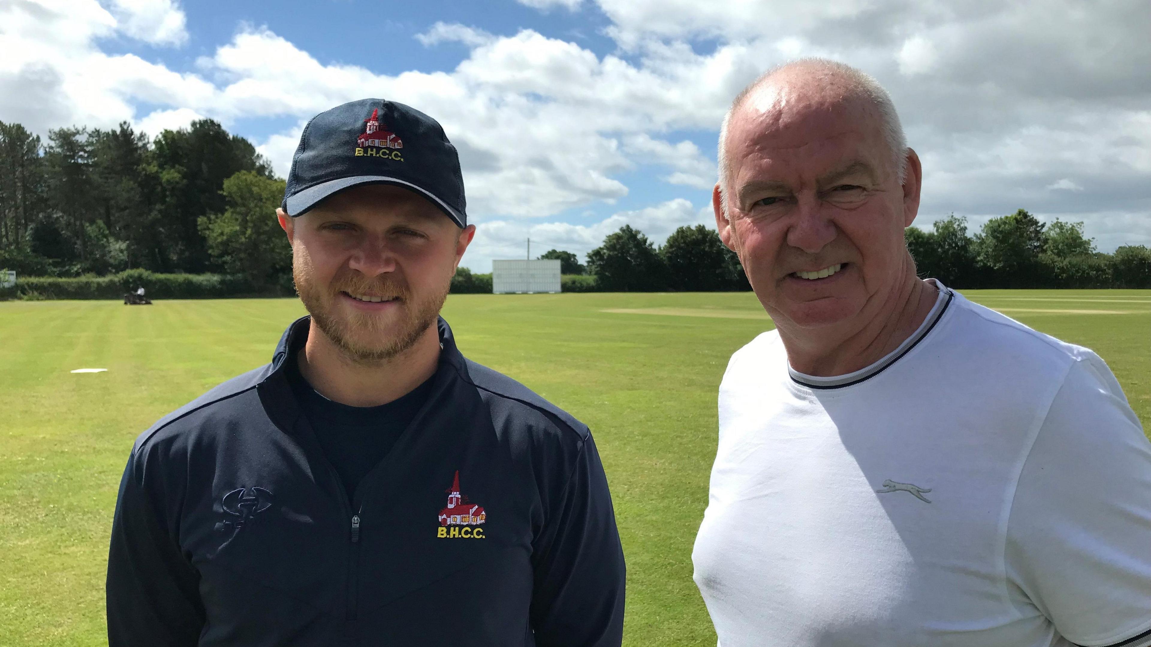 Harry Bolland in a blue club hat and top and Andy McIlroy in a white T-shirt, standing on the outfield at the club 