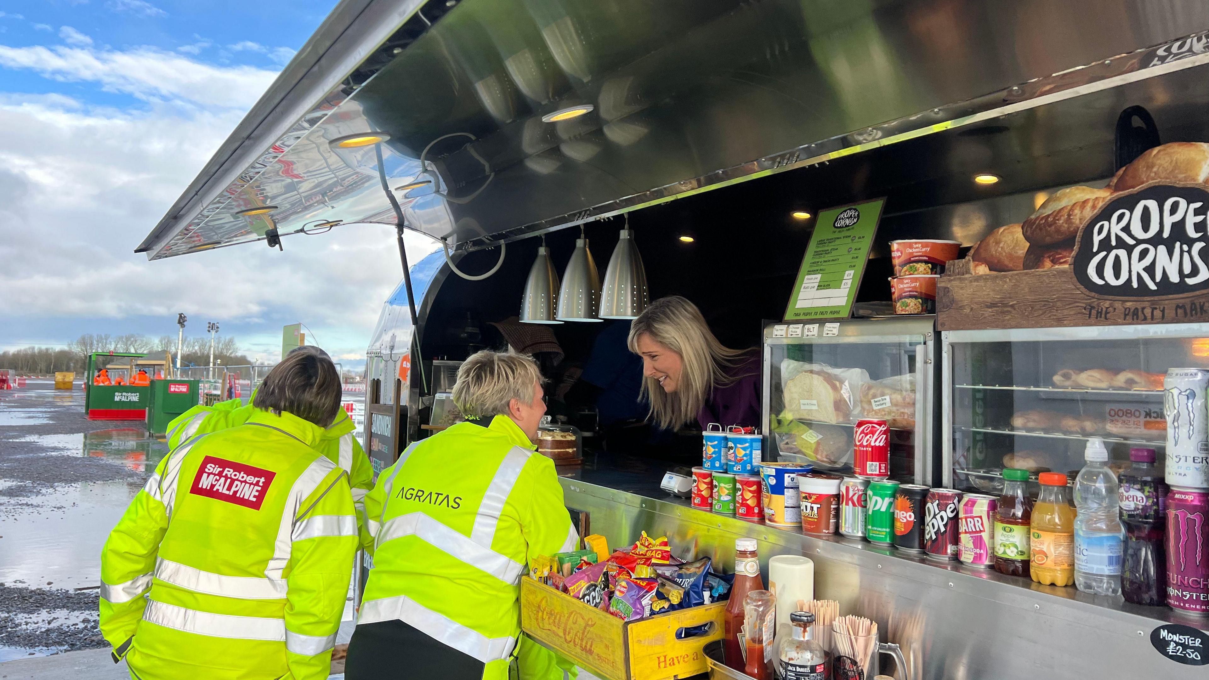 Three people in high-voz jackets and construction workers ordering food at the food van. There's food in the counter from drinks to pasties and toasties. There's a women leaning over taking their order.