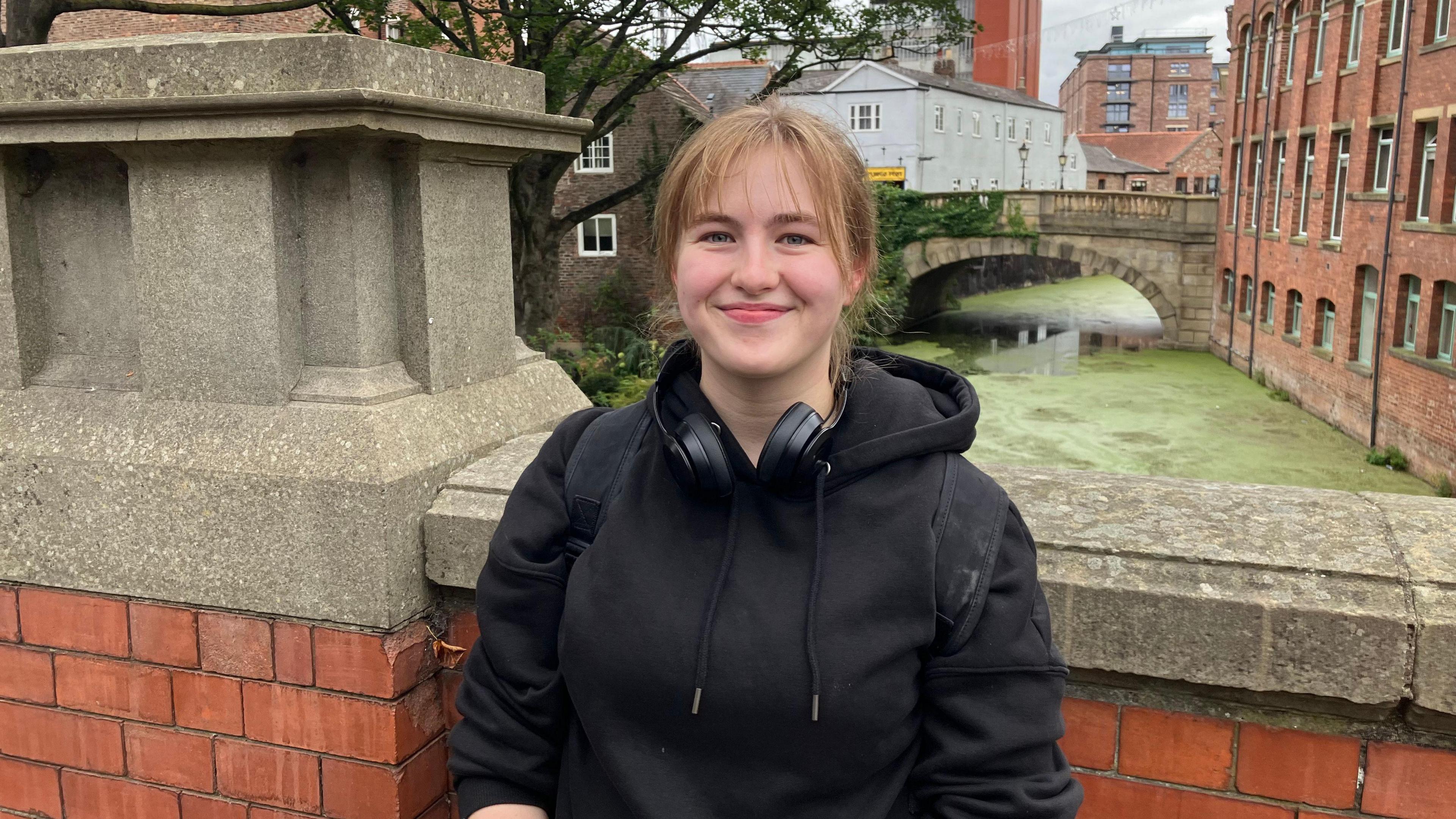 A young woman standing on a bridge above a green waterway covered in duckweed. She is smiling and wearing a black hoodie with black headphones.
On one side of the rivers is a garden with a tree and some shrubs and the other side has some brickwork and a small wall. Further down the river you can see a stone bridge.