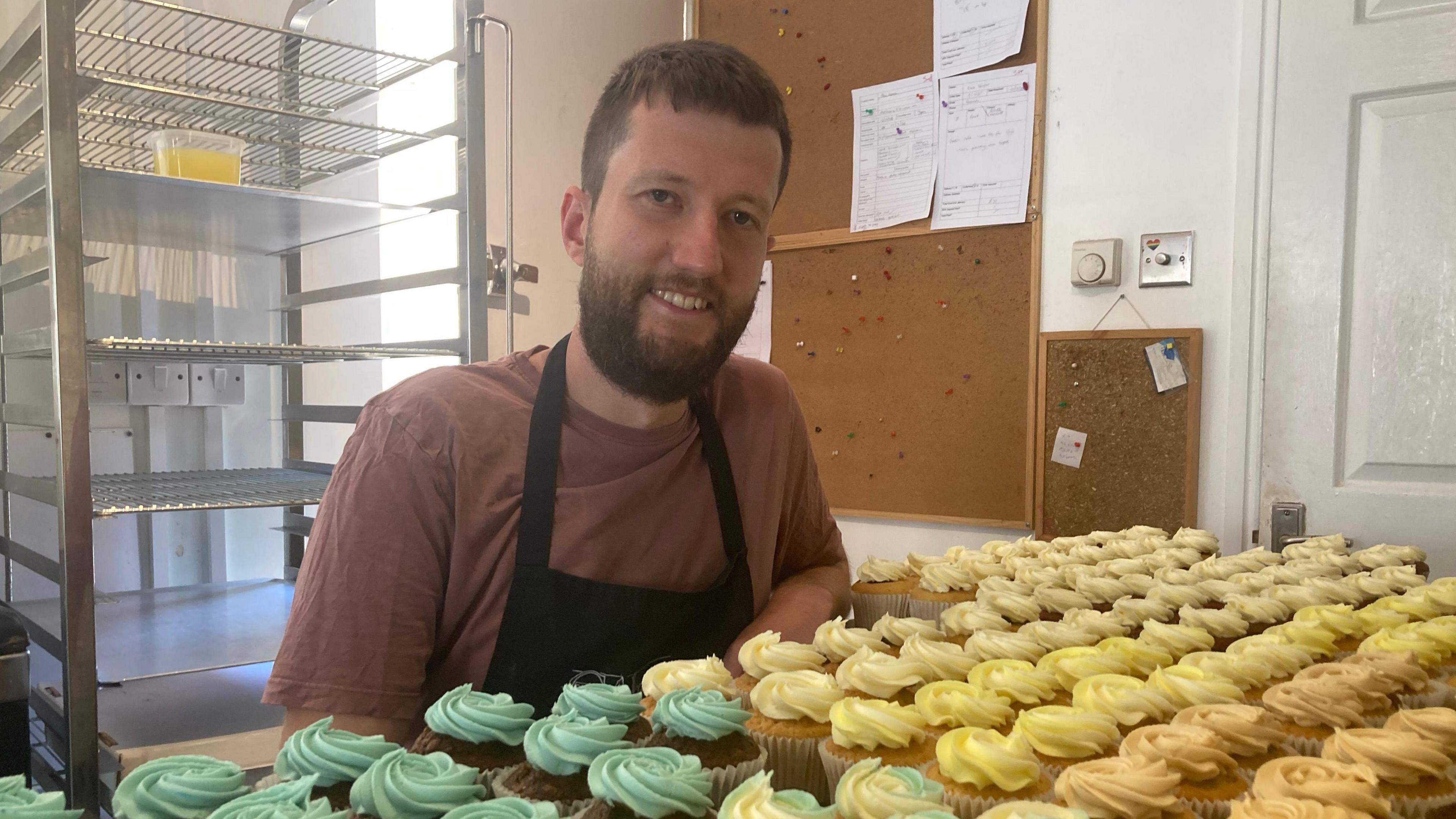 A man with a beard stand behind a large tray of cupcakes, decorated with green and yellow icing.