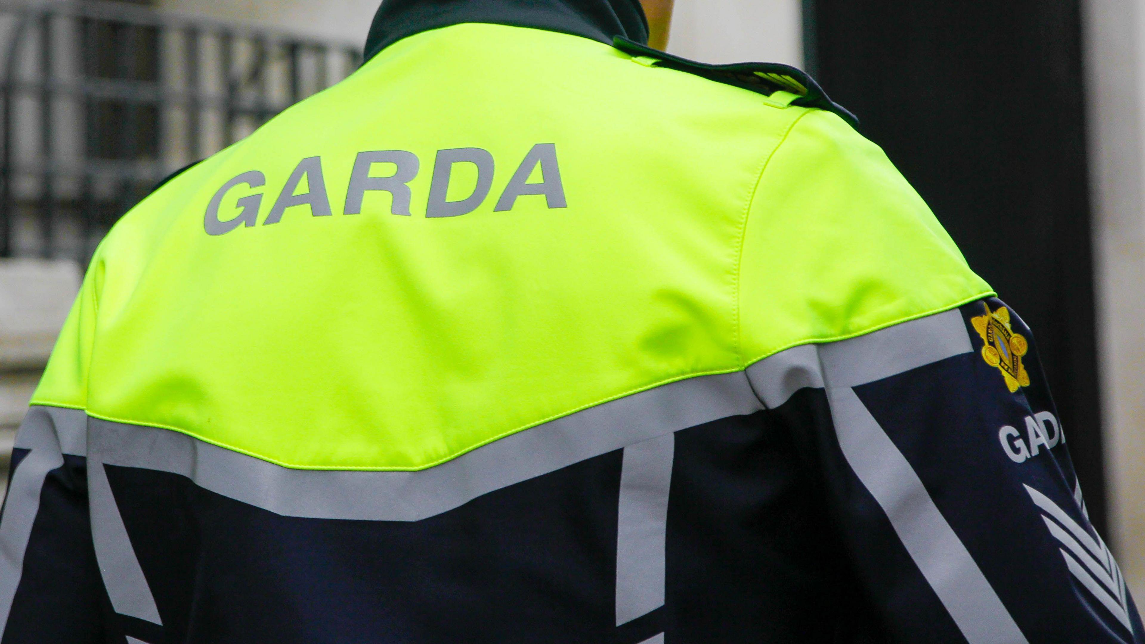 The back of a Garda officer. Garda is written on the luminescent top of his uniform the rest is navy with grey trimmings. The background is out of focus and his head is not visible. 
