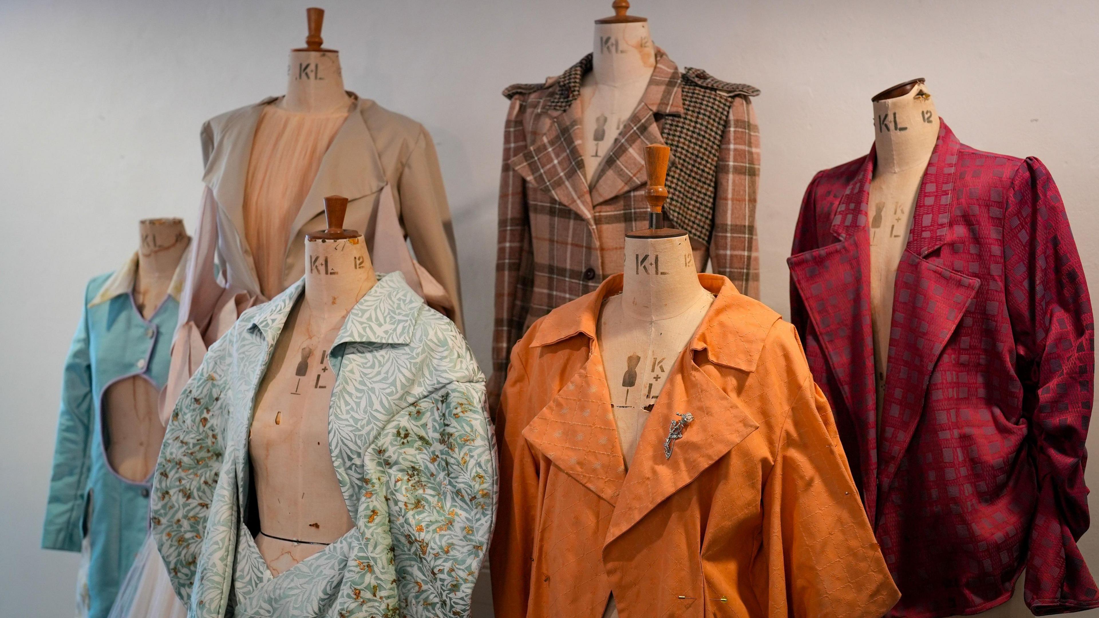 Six different coats displayed on headless wooden mannequins. There is a rich red cat with a large collar, an orange trench coat, a beige tartan coat, a beige trench coat, a blue floral coat and an aqua-coloured coat