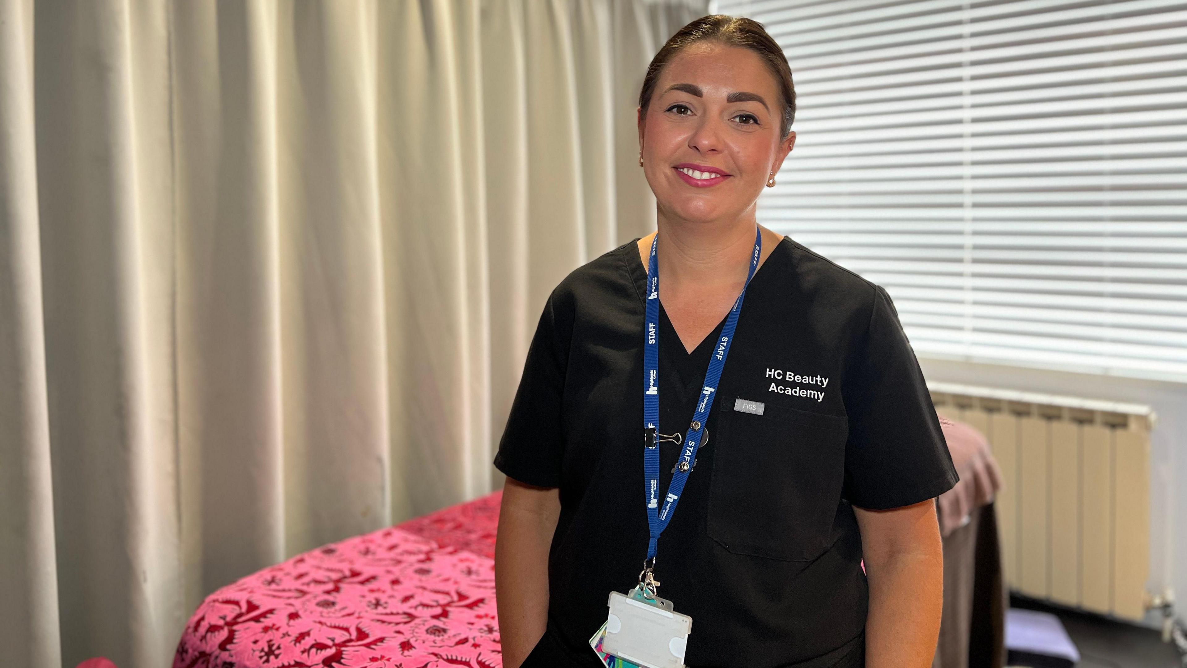 Kayleigh smiles at the camera as she stands in front of a massage bed and a drawn curtain