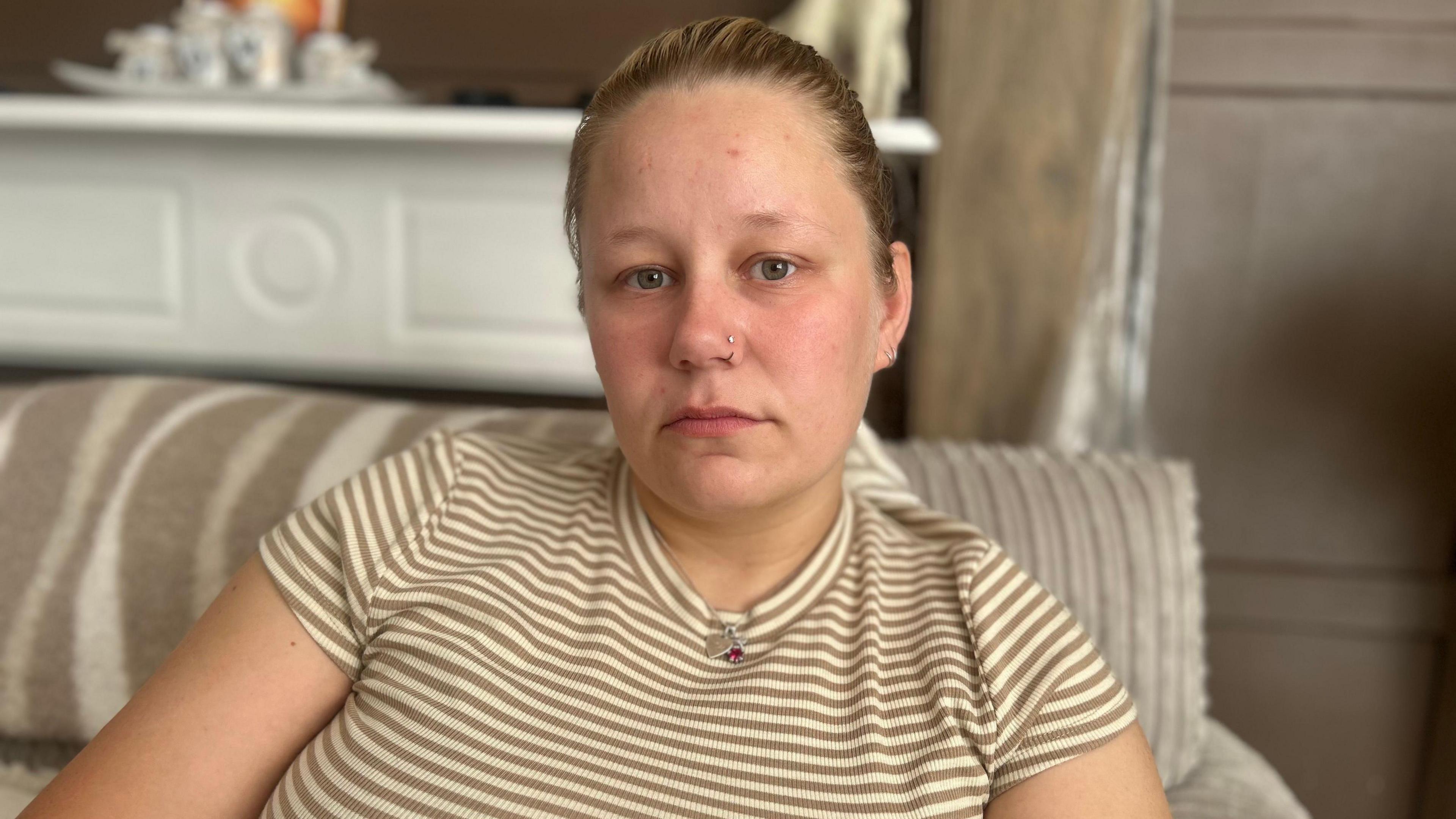 A person sits on a striped sofa in a living room, facing the camera. The person is wearing a short-sleeved beige and white striped top, a necklace, and a nose stud. Tattoos are visible on both forearms. The background includes a white mantelpiece and wooden wall panels, with a shallow depth of field keeping the person in focus while the room behind is softly blurred.