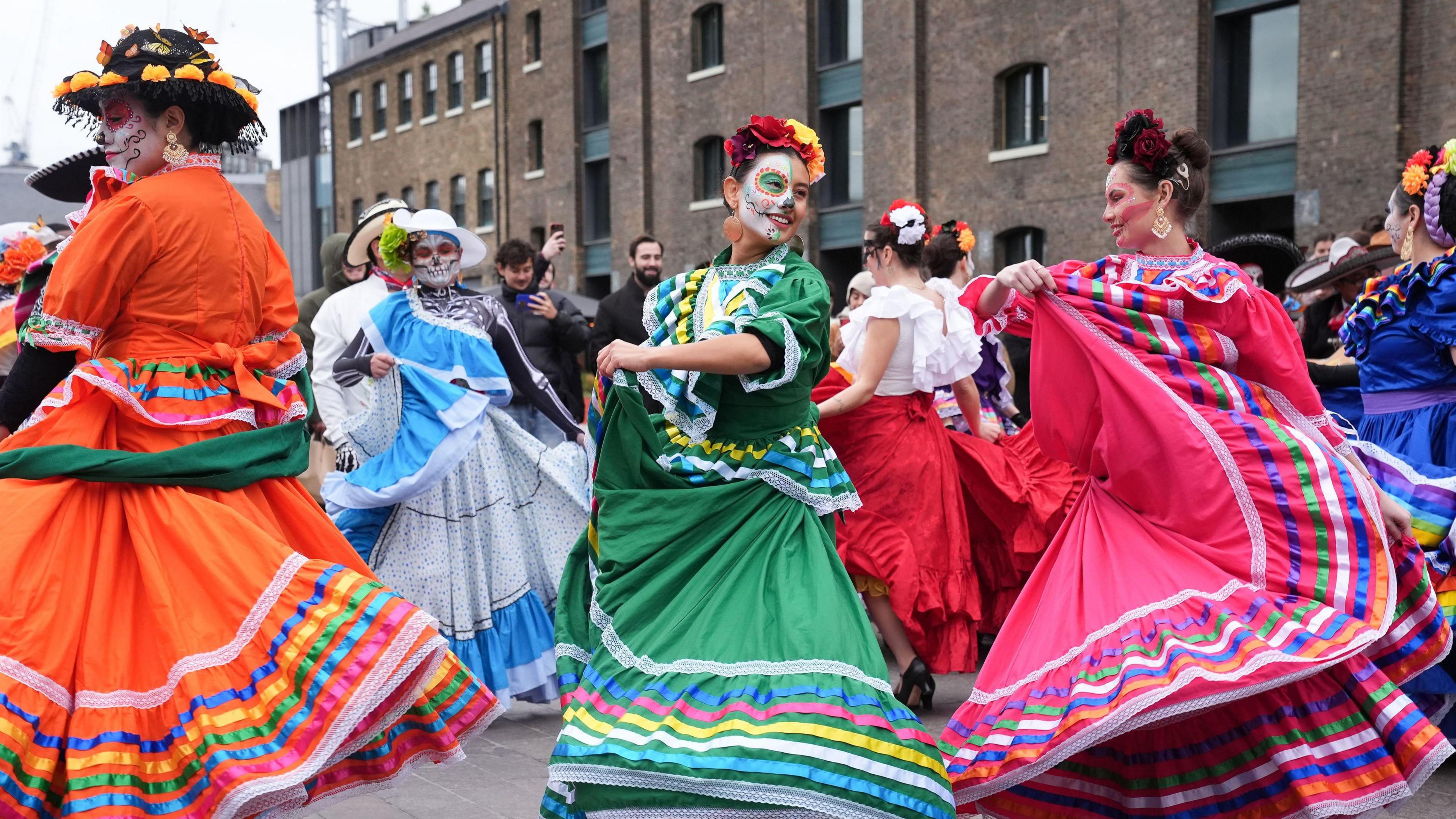 Women wearing colourful dresses dancing in the street. They have their faces painted as skulls.