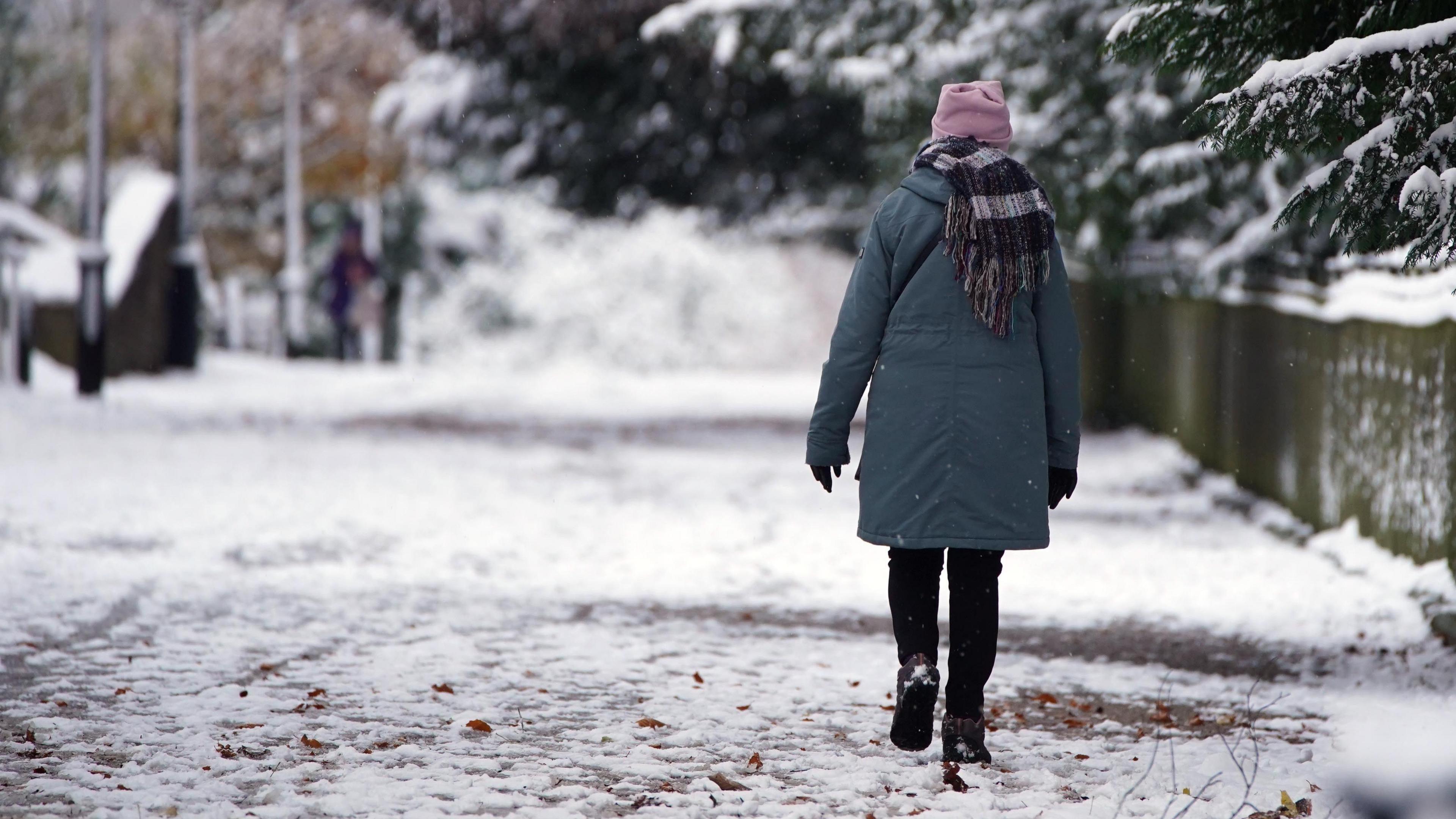 A woman wrapped up in a winter coat, scarf, hat and gloves walks along a snowy pavement