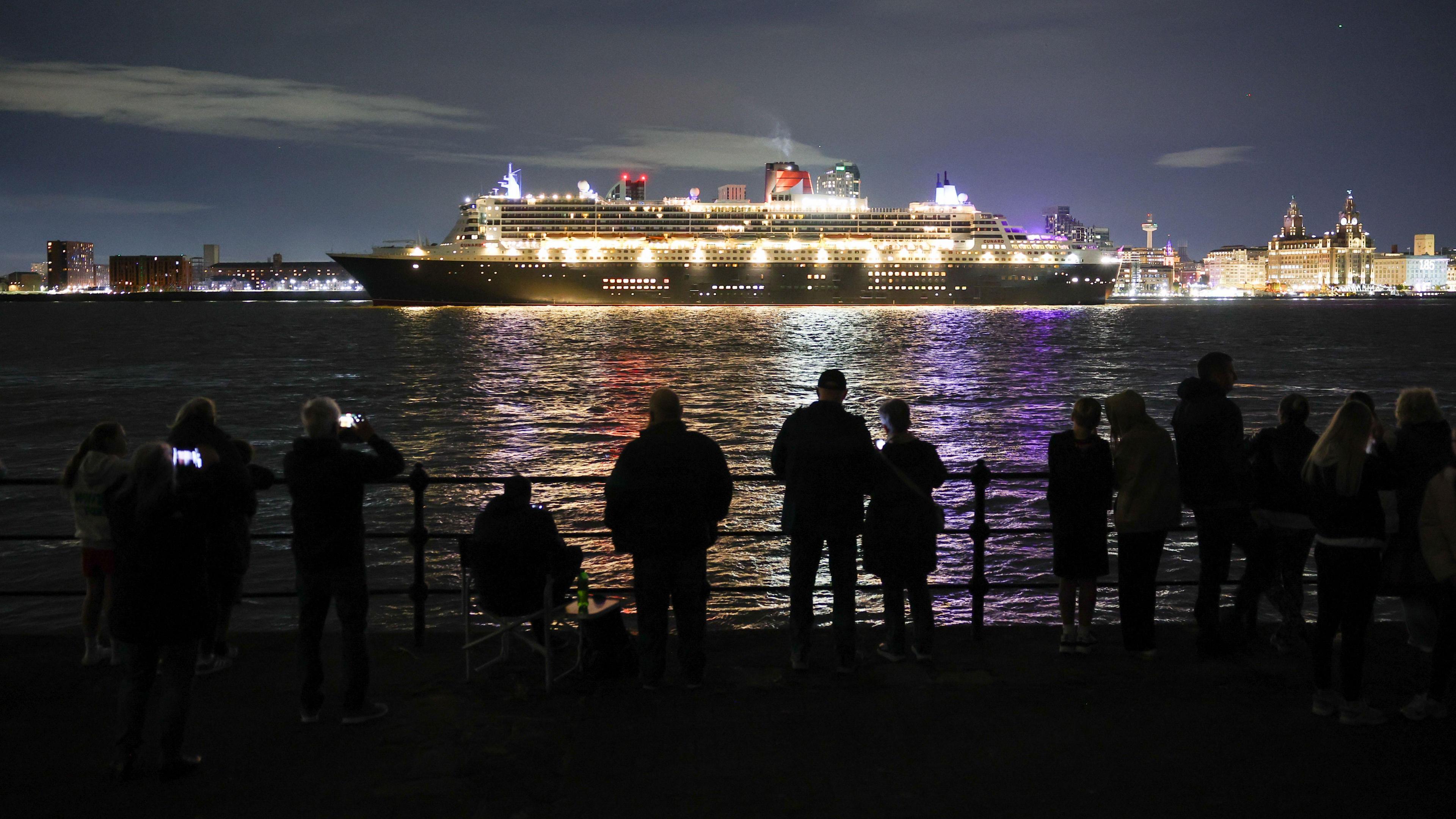 More than a dozen people stand and sit on the Wirral side as they watch the illuminated liner sail past on the river with Liverpool in the background