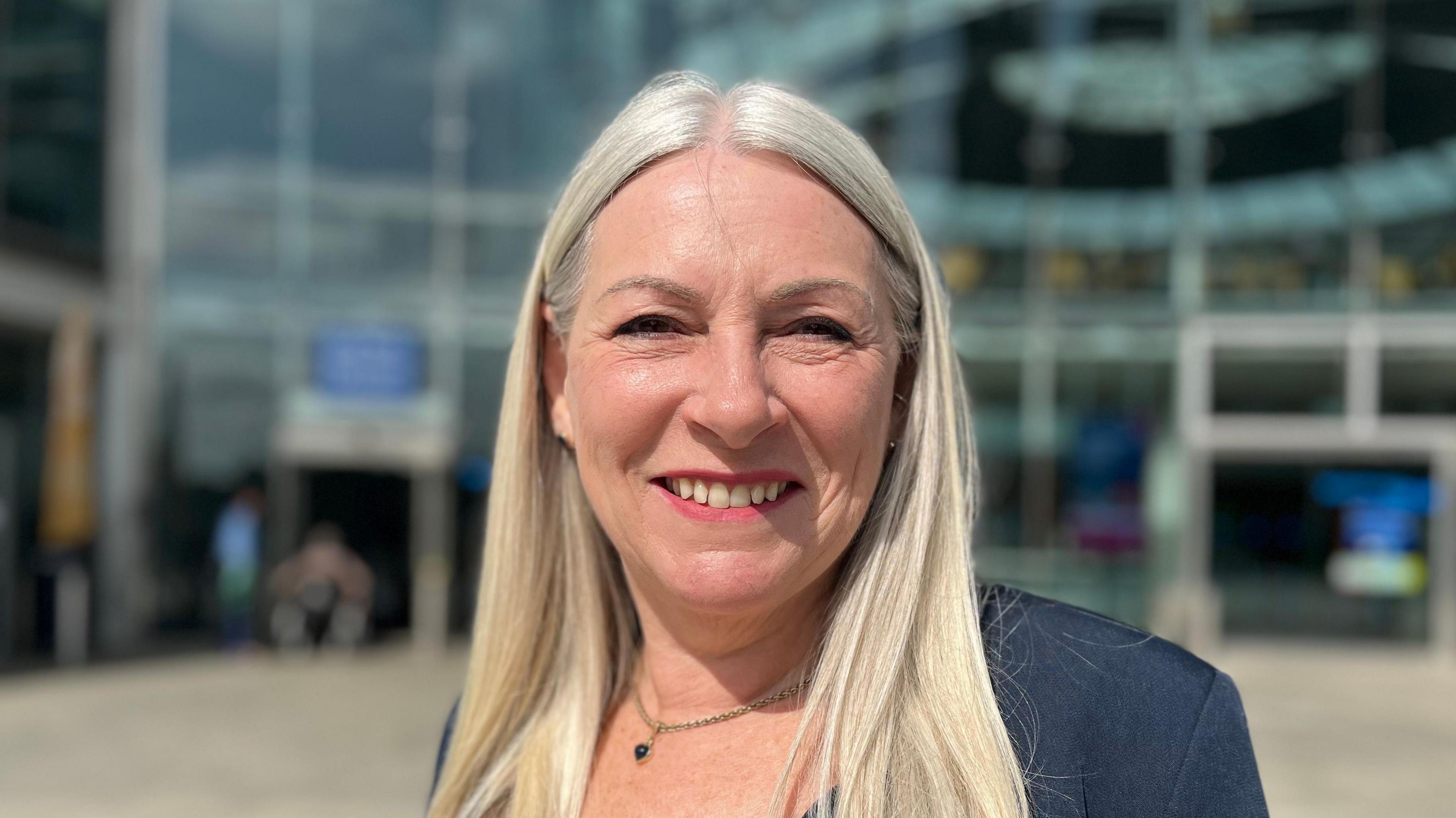 Kay Mason Billig is smiling at the camera whilst standing outside the Forum in Norwich. She's wearing a navy blue suit jacket.