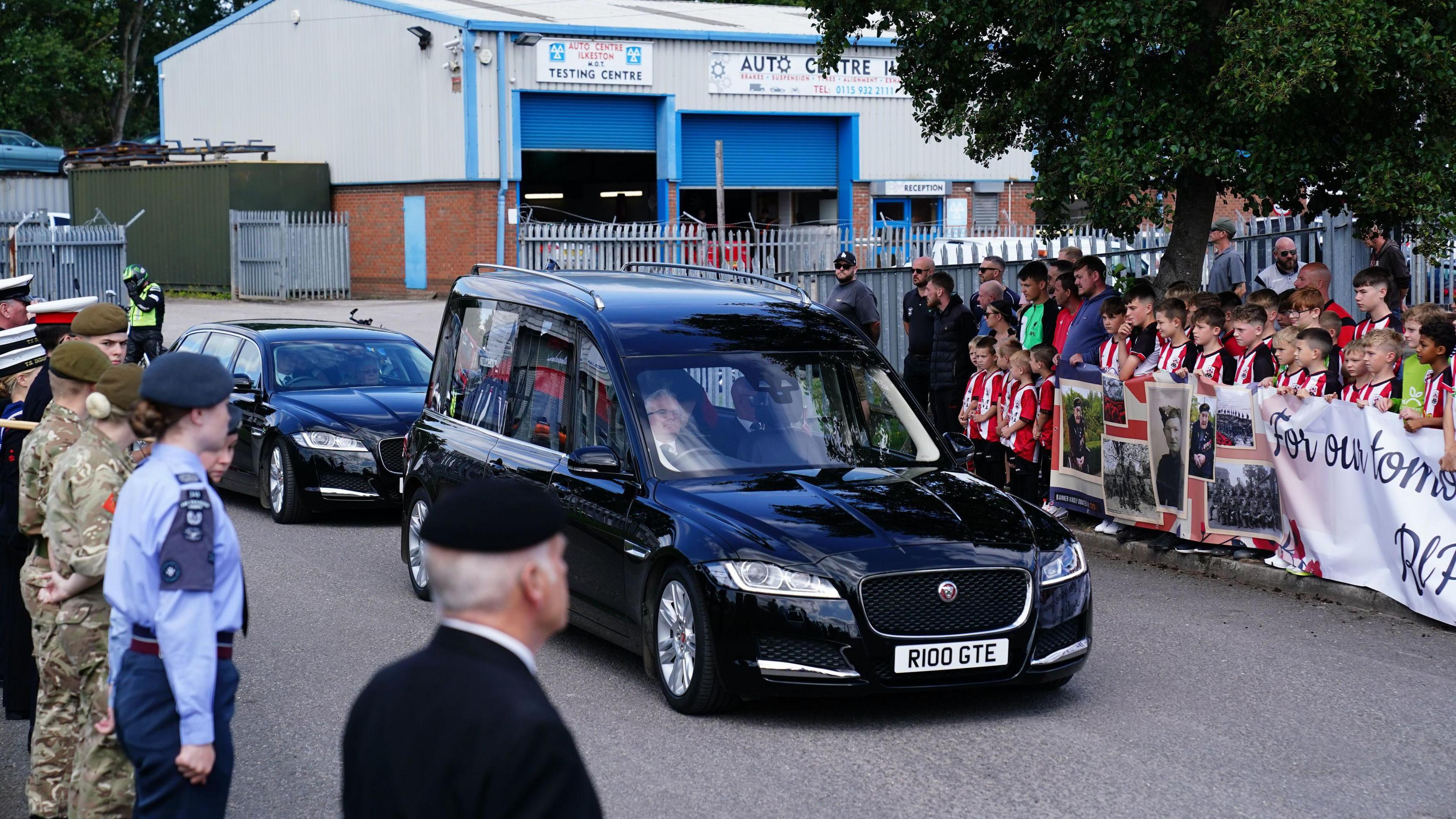 A children's football team and people in military uniform line the streets while a hearse travels along the road
