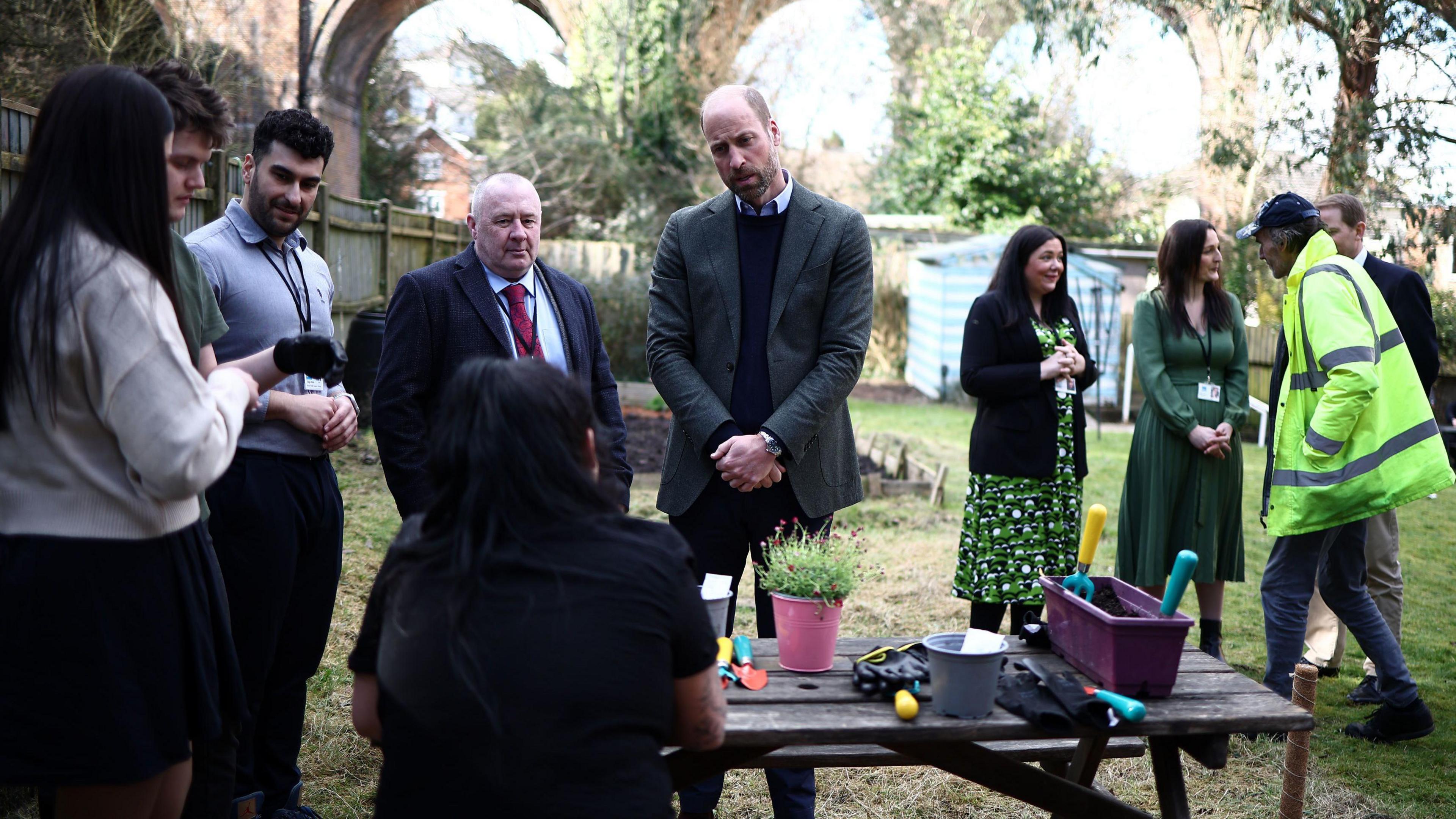 The Prince of Wales talking to residents in the garden of Millennium House. He is standing in front of a table where there are gardening tools and planters. There are nine other people in the image, including officials, reporters and residents.