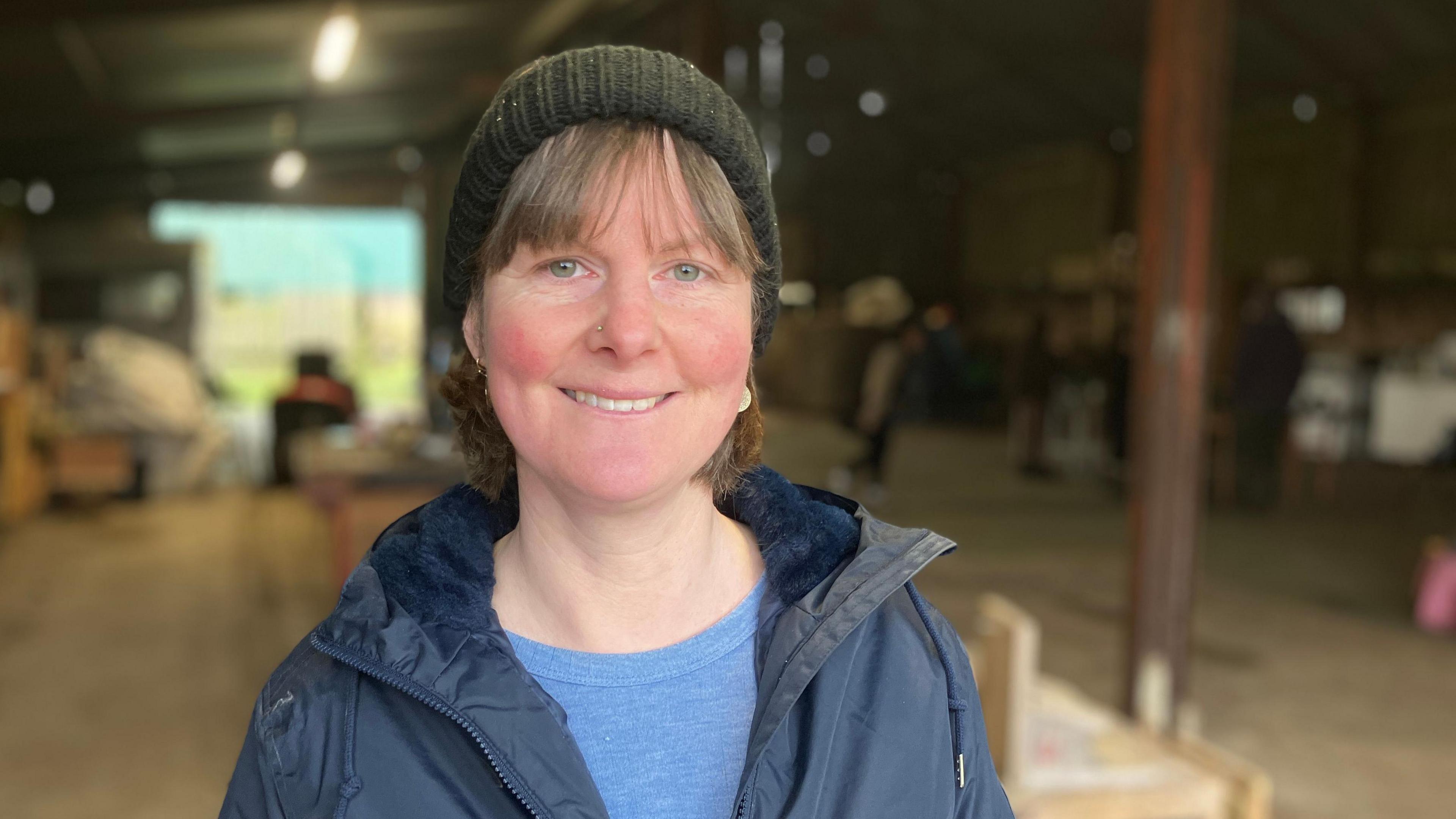 Jill Heslop is standing in a large barn out of focus behind her, and is wearing a dark green woollen hat and dark blue waterproof coat, open at the top to reveal a pale blue T-shirt. She has short, light brown hair with a fringe and is smiling.