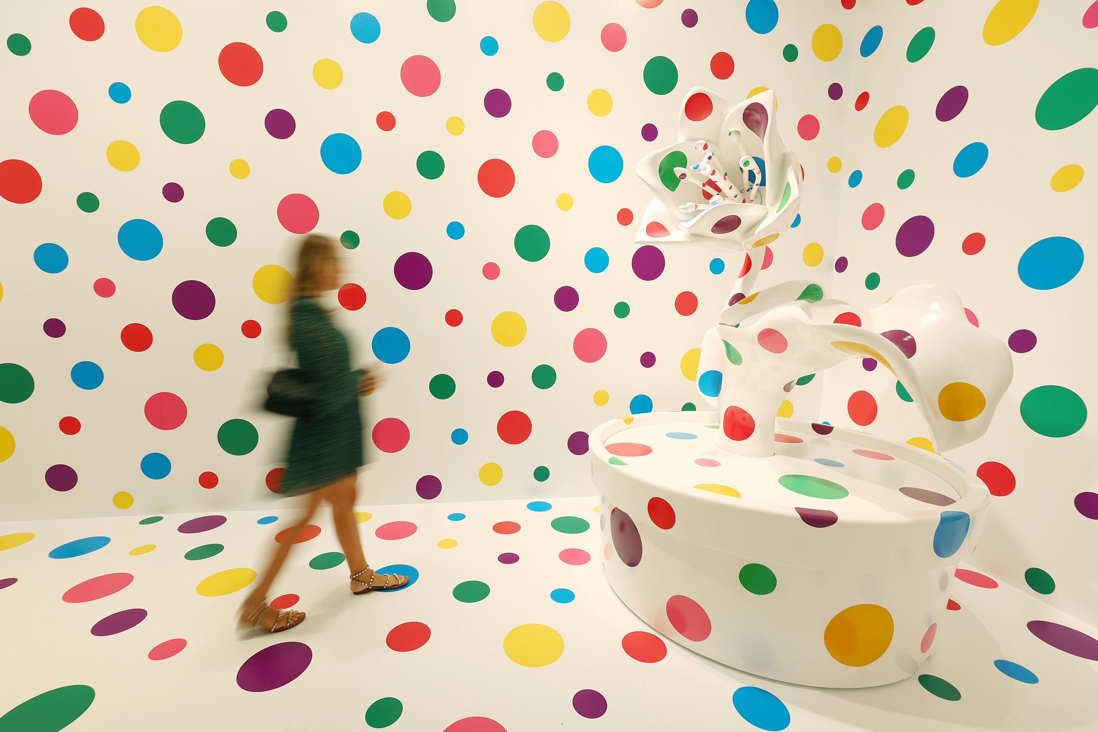 A blurred gallery visitor in front of a white wall and sculpture covered in multi-coloured spots