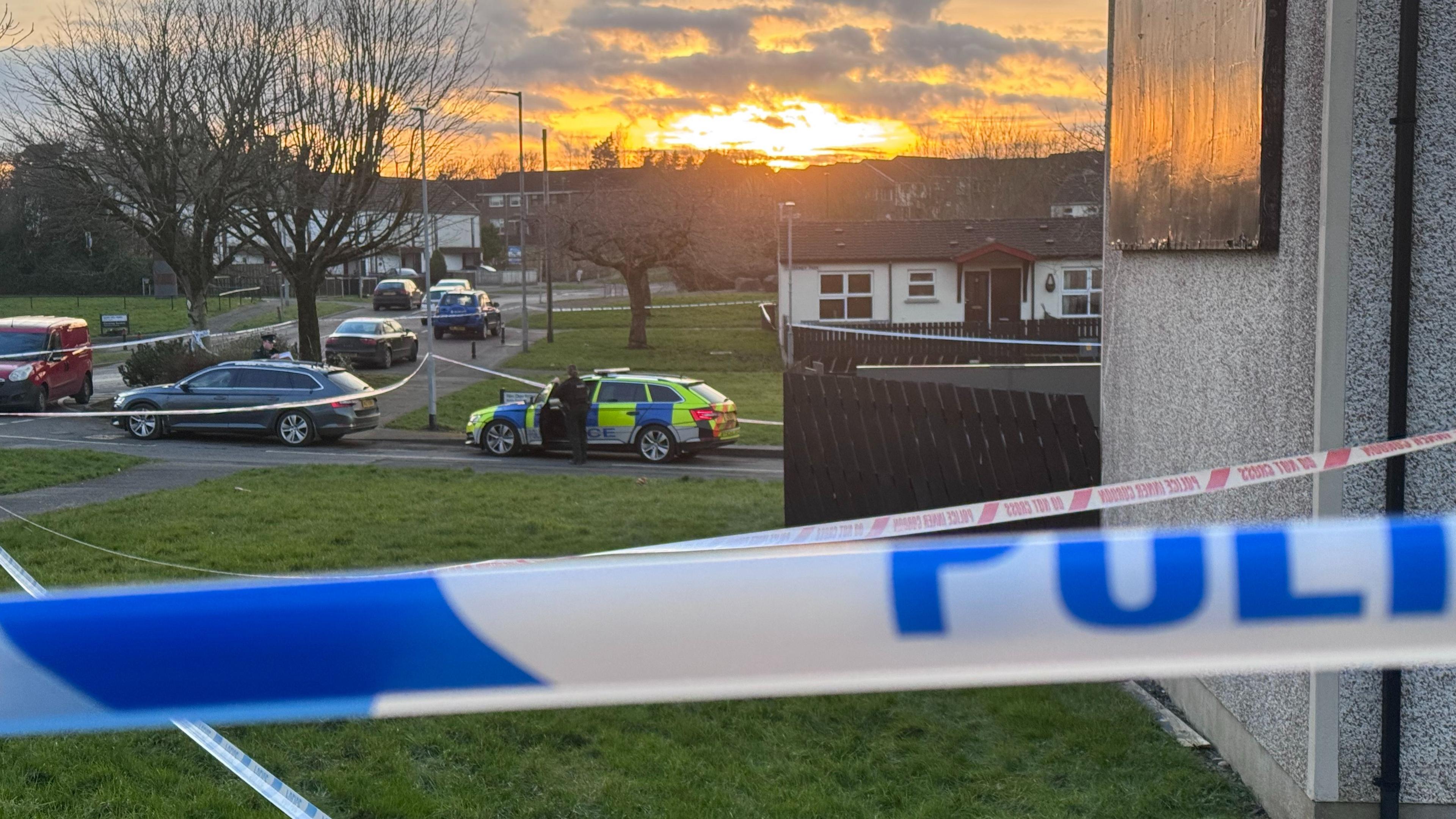 Blue and white police tape is cordoning off a street. It's dusk and the sunrise is in the distance.