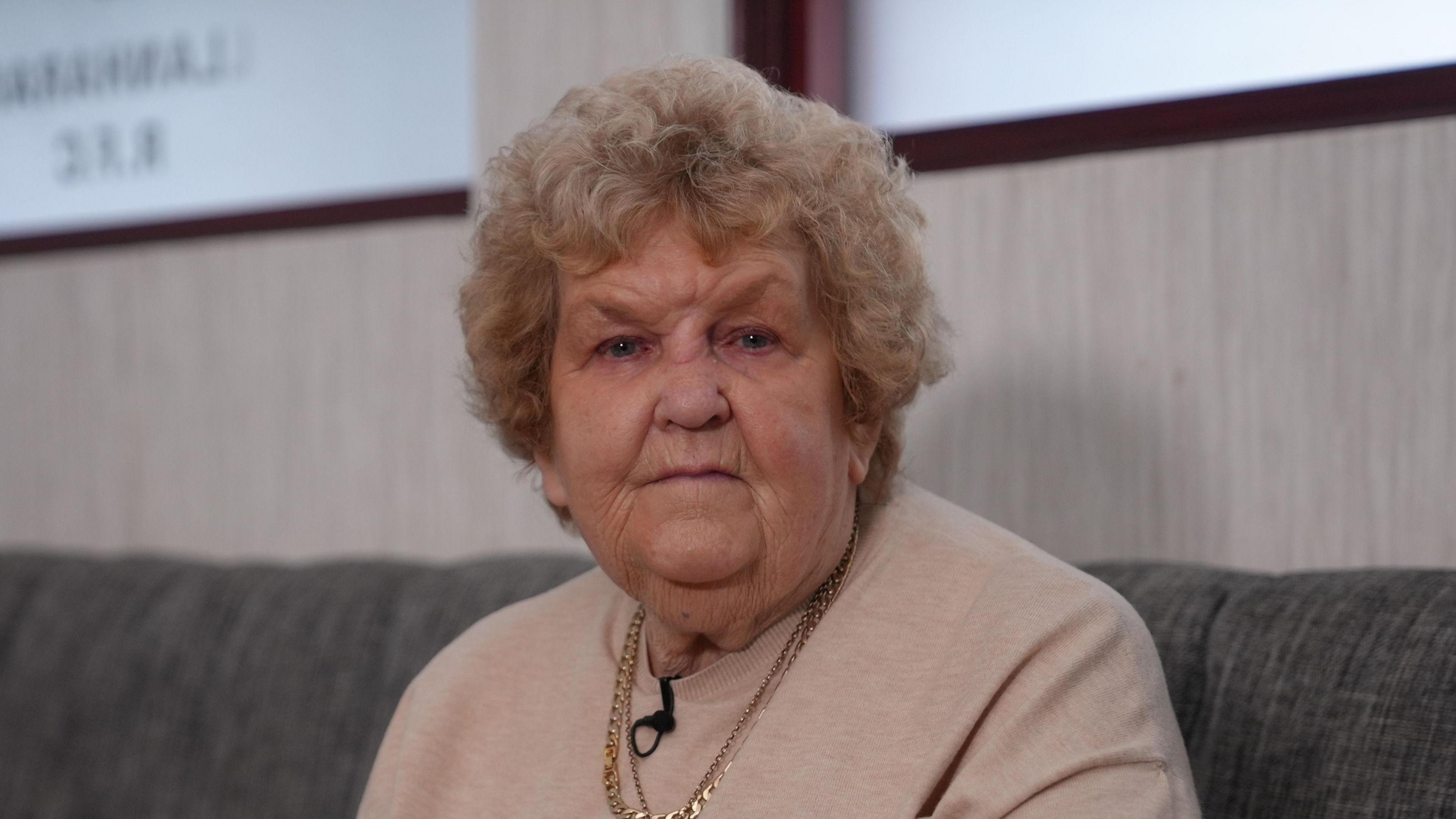 Woman in pale pink sweater wearing a gold chain necklace and short, curly blonde hair, looks at the camera