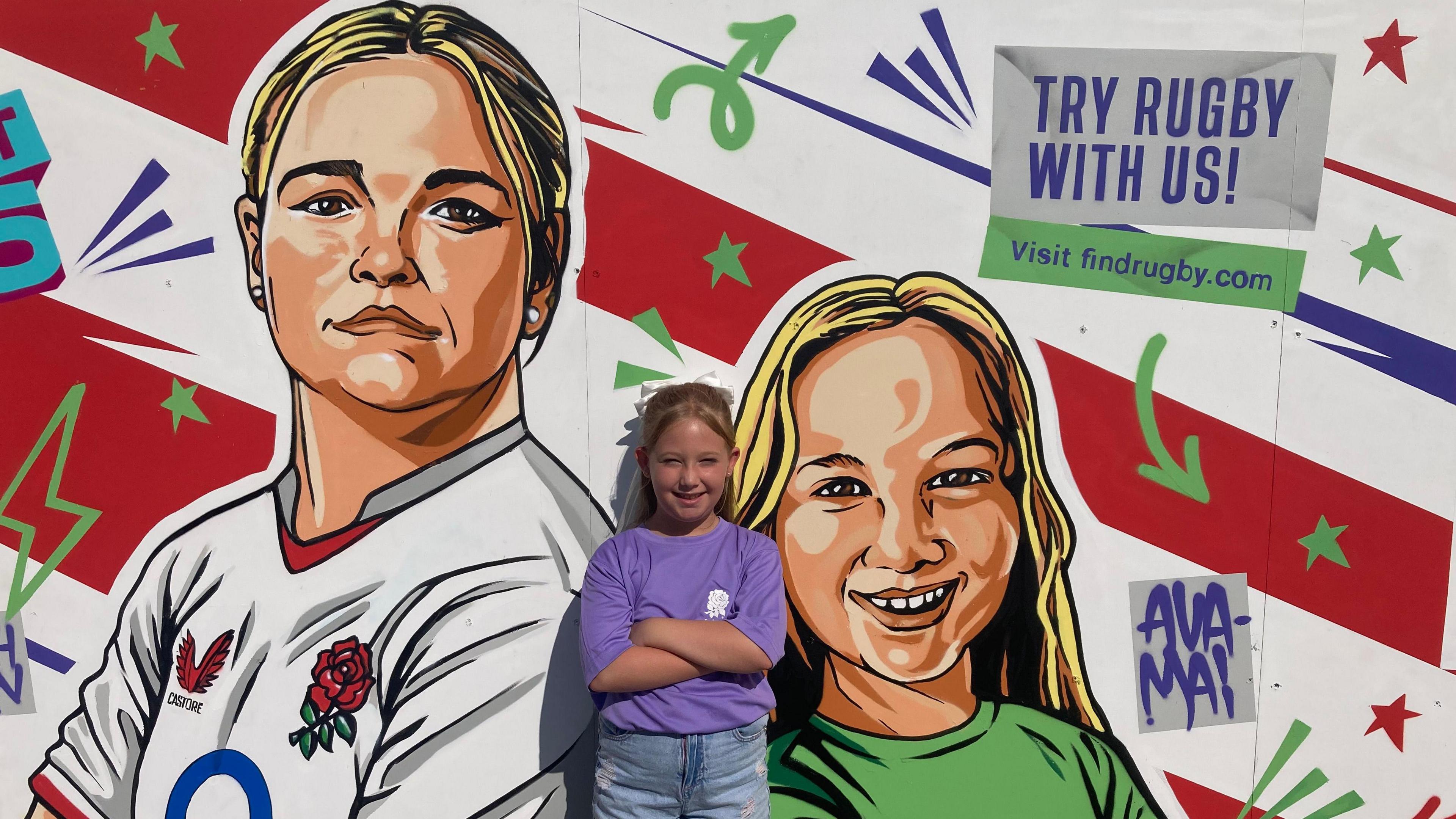 A painted mural depicting England rugby player Sarah Bern and nine year old Ava-Mai smiling and looking at the artist with red and white stripes in the background and colourful arrows and stars on it. The real Ava -Mai stands with her arms folded in front of it, she is smiling at the camera and wearing a purple t shirt and denim shorts
