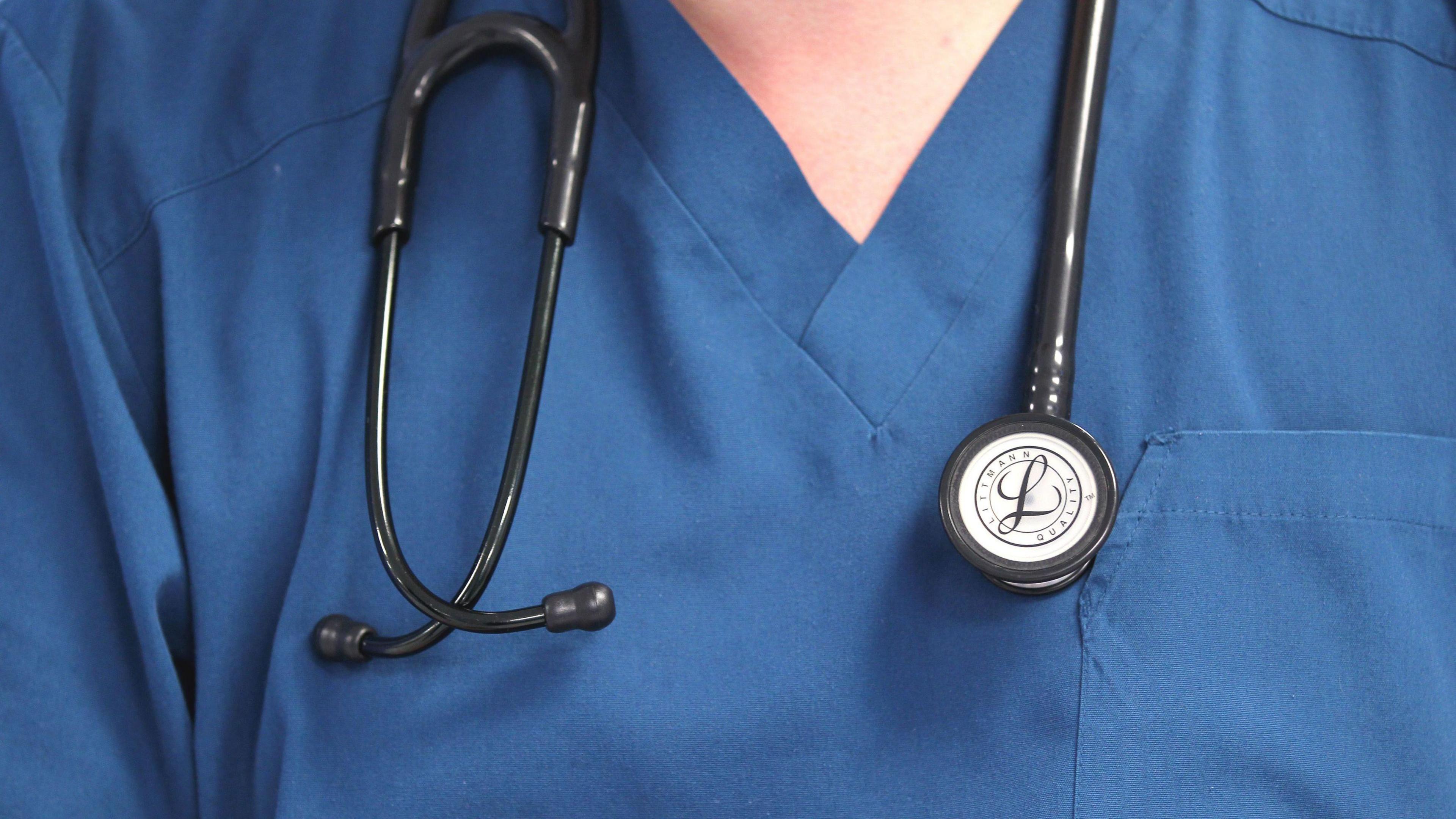 The upper chest of a doctor wearing sky blue scrubs with a back stethoscope draped around the neck.