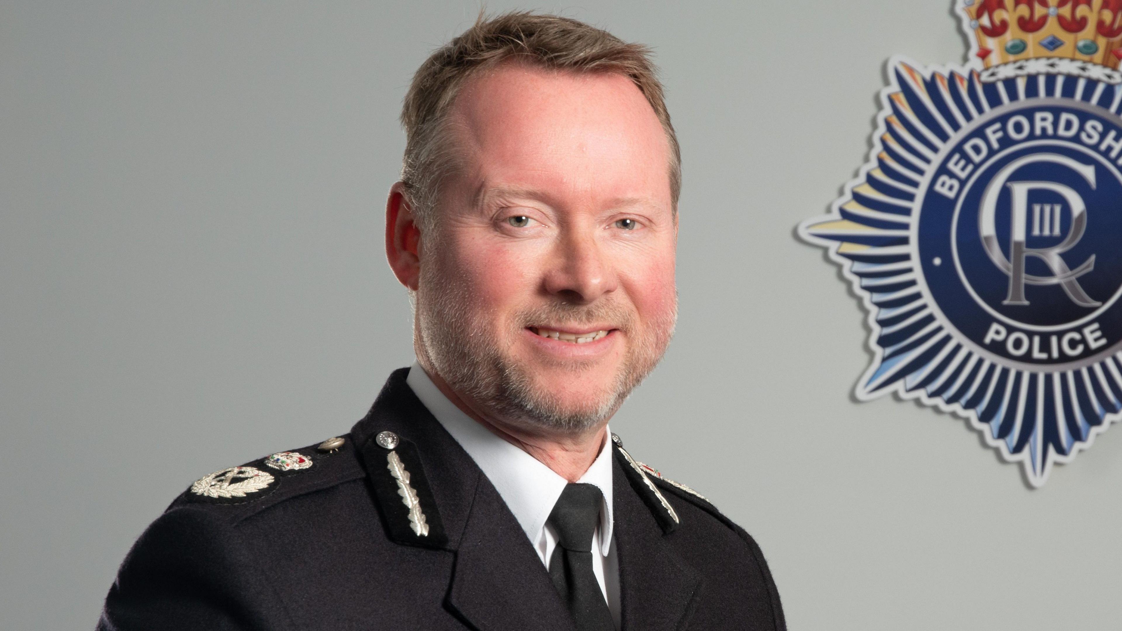 Chief Constable Trevor Rodenhurst is wearing a police uniform and smiling. Behind him on the white wall is a badge for Bedfordshire Police