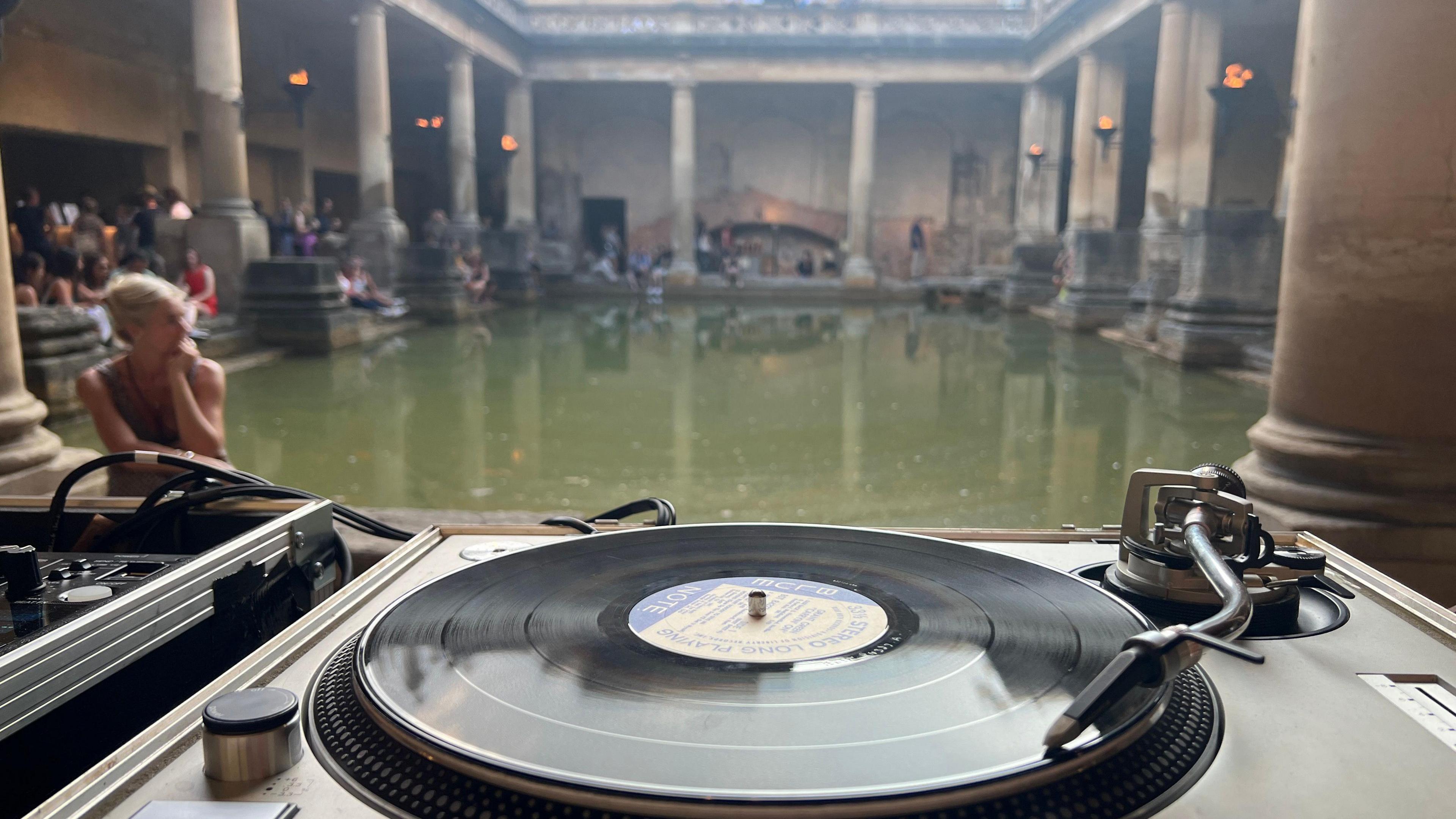 A deck with a vinyl playing is in the foreground. In the background is a Roman thermal bath, which looks like an open-top green rectangular pool, with stone pillars surrounding it. There are people sitting around the pool. 