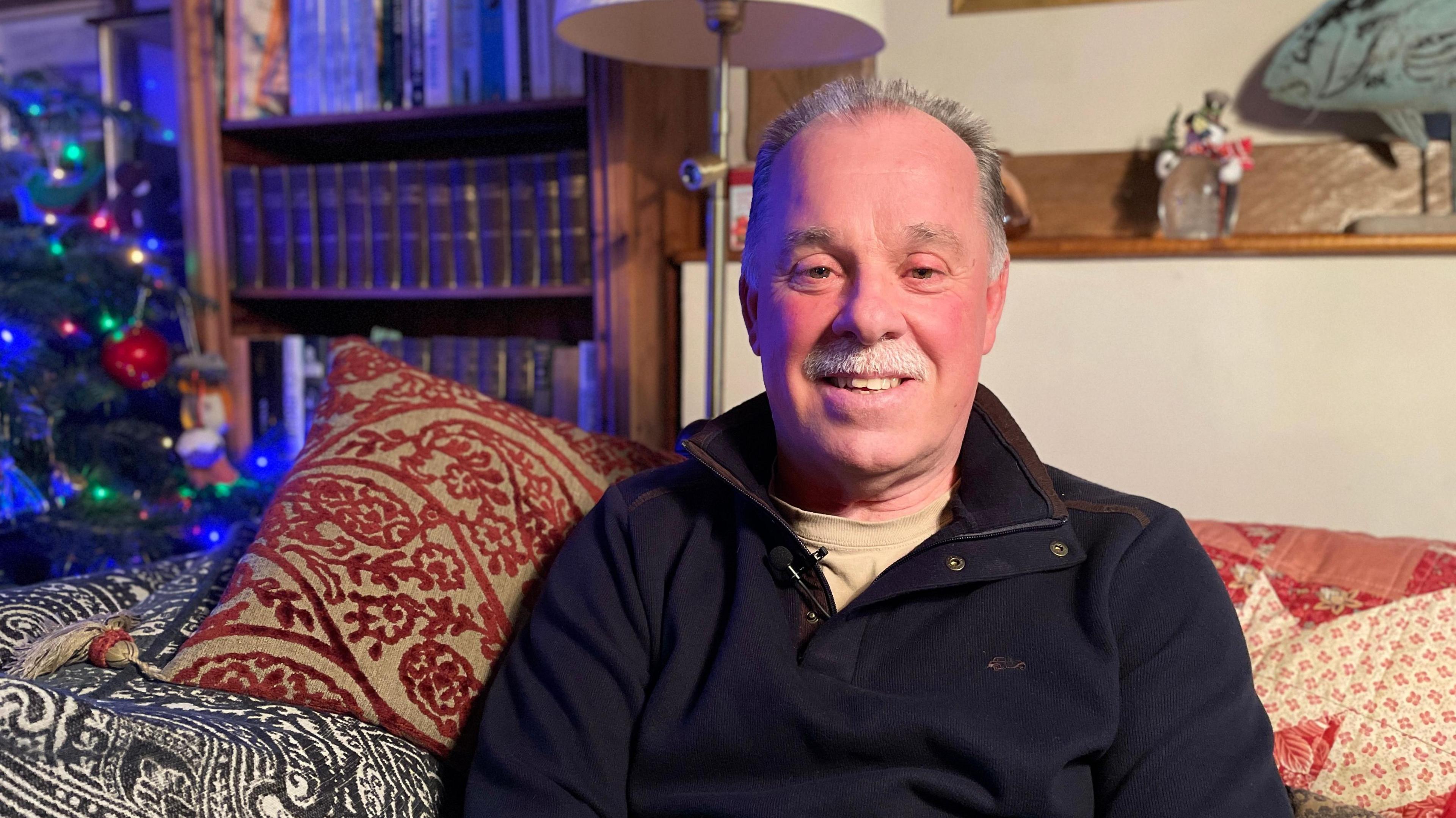 David Llewellin at his home in Monkland, Herefordshire. He is sitting on a sofa, on his left is a Christmas tree and behind him is a bookcase and a shelf with ornaments on it.