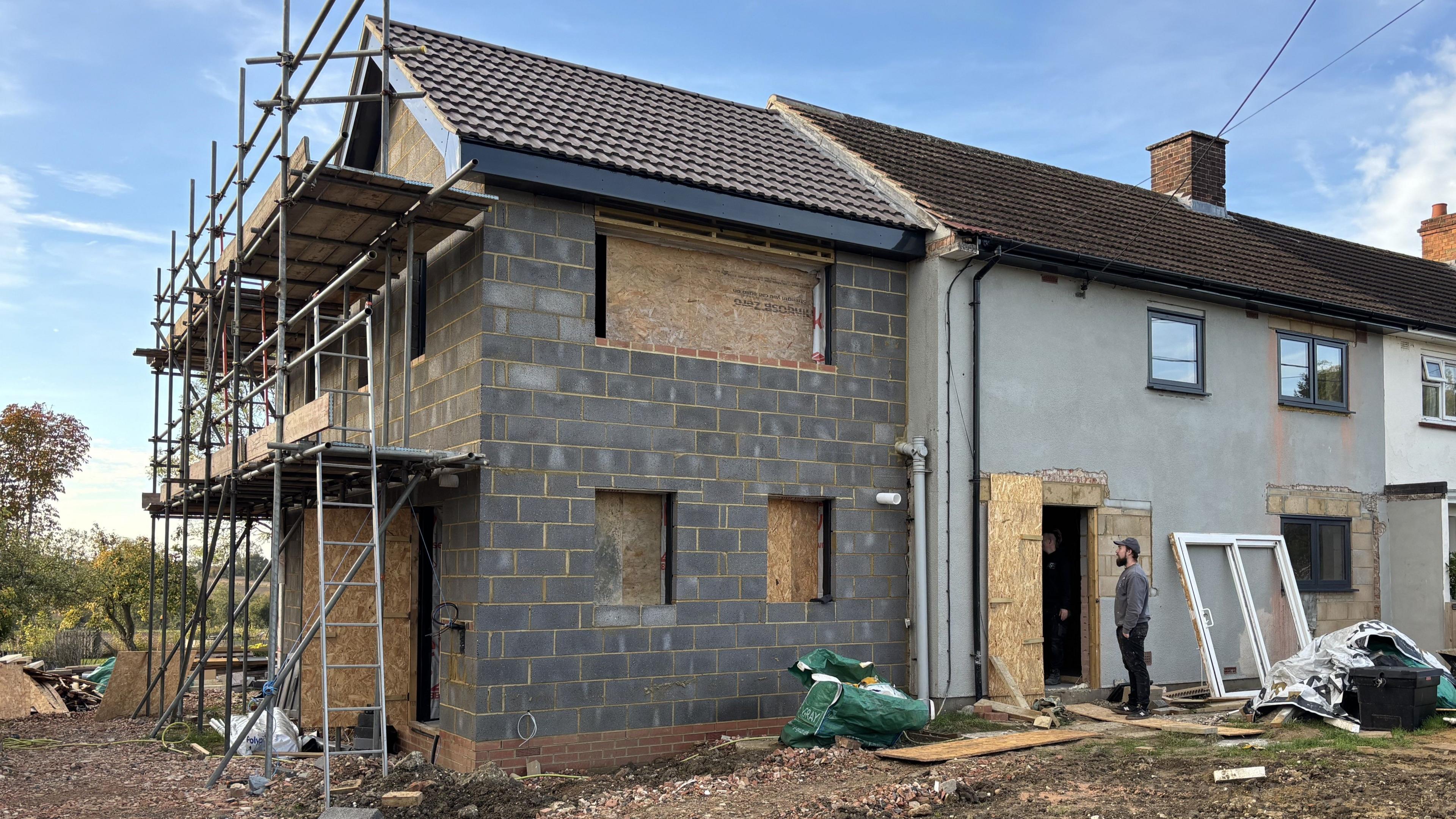A photo taken outside where a house renovation is taking place. A new house build can be seen attached to already constructed homes. It is made of large grey bricks and has scaffolding on one side of it. The windows are covered in wooden planks. A man stands in the doorframe of part of the house. 