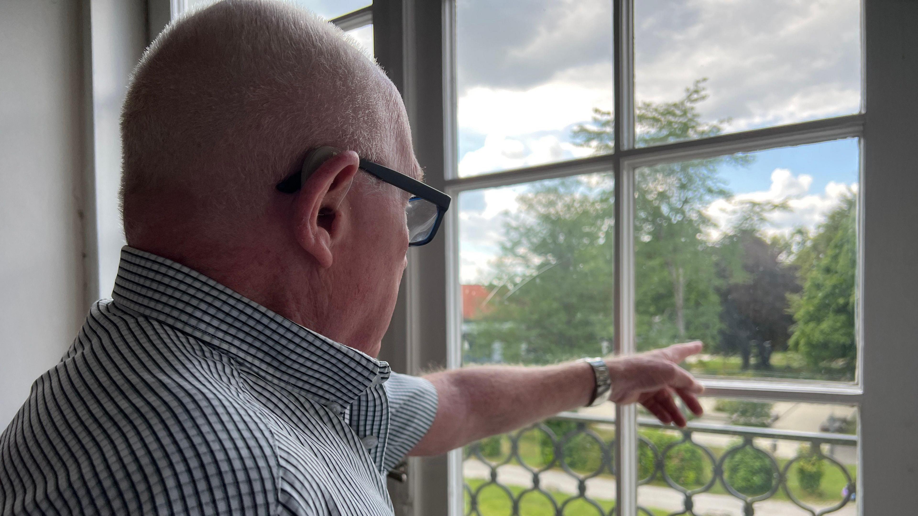 Roy has short hair and glasses, and is wearing a striped shirt, stands indoors and points outside while looking through a window. Outside the window, there is a green lawn, trees, and a partly cloudy sky."