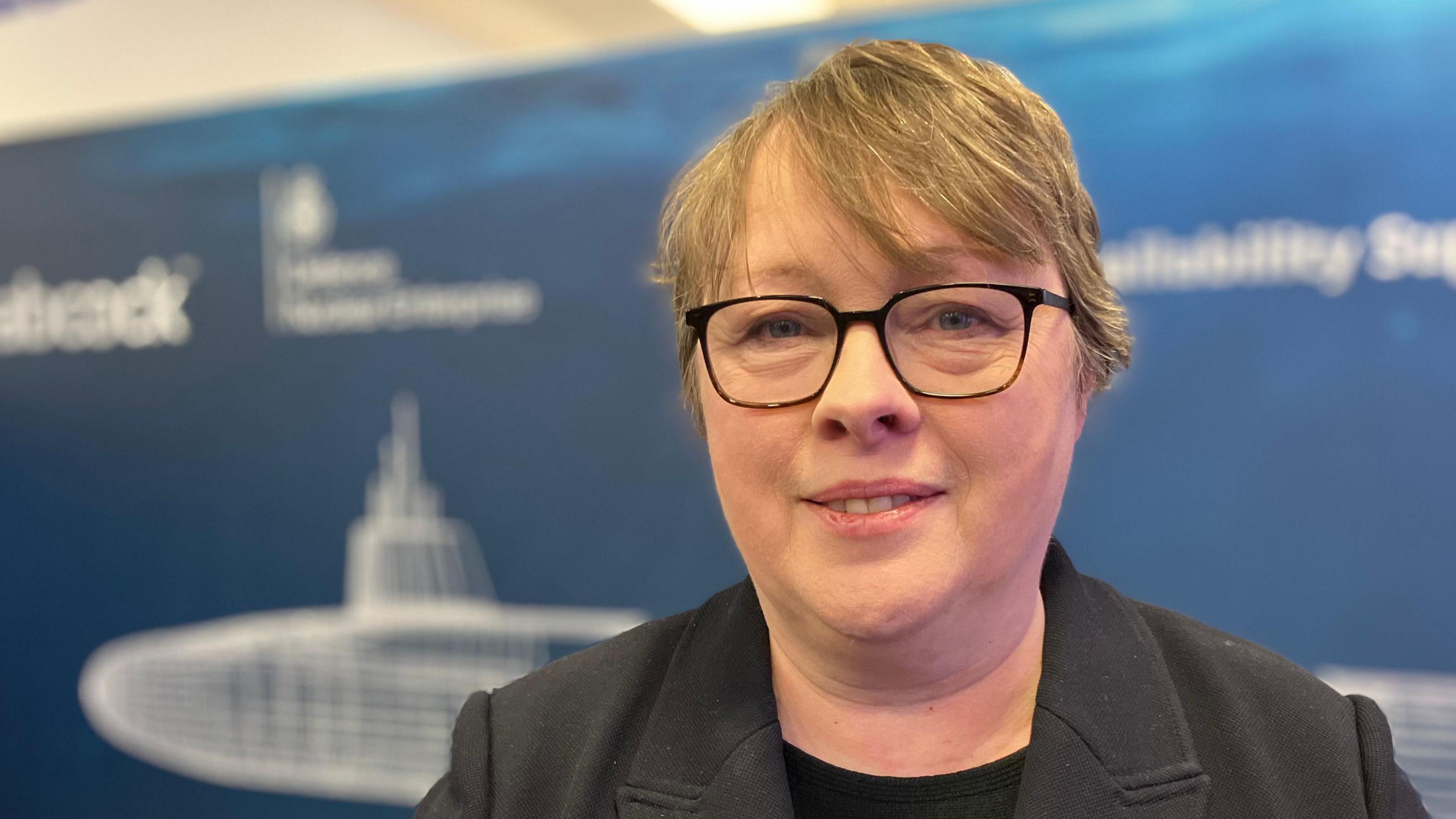A head shot of former defence procurement minister, Maria Eagle MP. She is wearing glasses and a black jacket over a black T-shirt.
