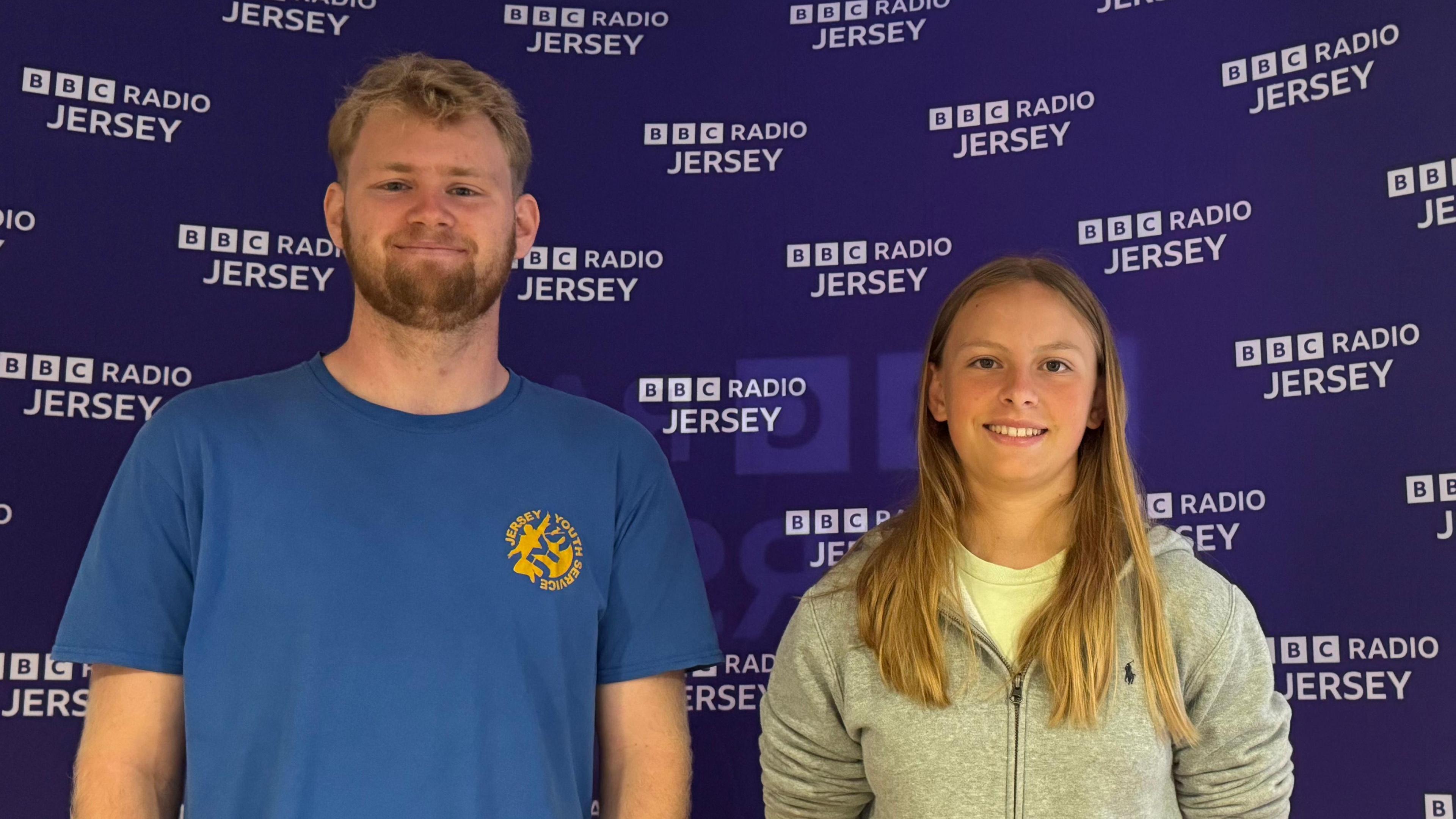 Josh Twohig-Jones on the left wearing a blue top with a Jersey Youth Service badge, and Lauren wearing a grey hooded jacket. They are both smiling at the camera and stood in front of a purple Radio Jersey sign.