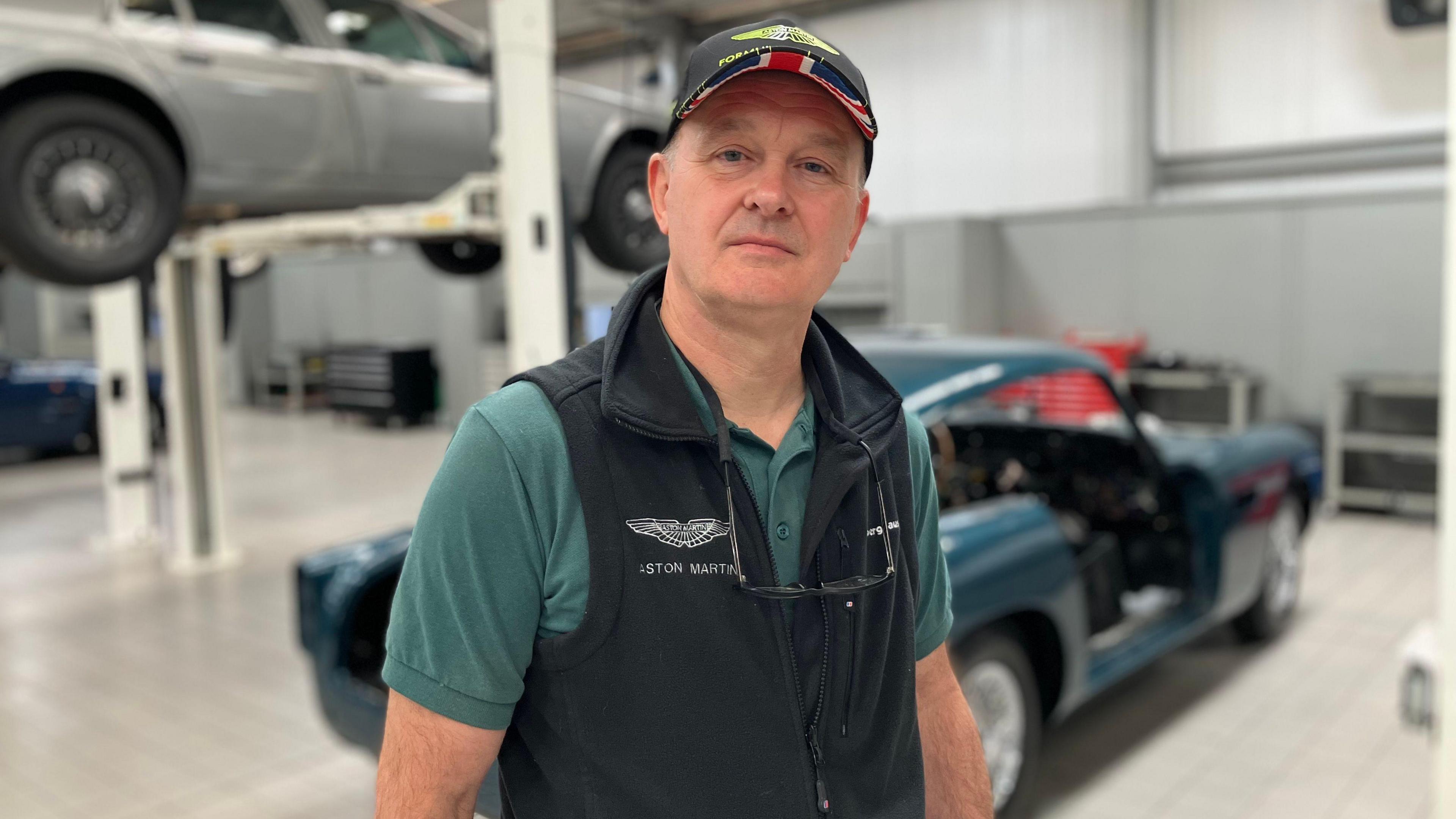 An upper body picture of David Alderman who is wearing Aston Martin branded clothing and a peaked cap. He has glasses hanging around his neck. The background is blurred, but two sports cars can be seen behind him. A blue one is on the floor. A silver one is hoisted up above head height. 