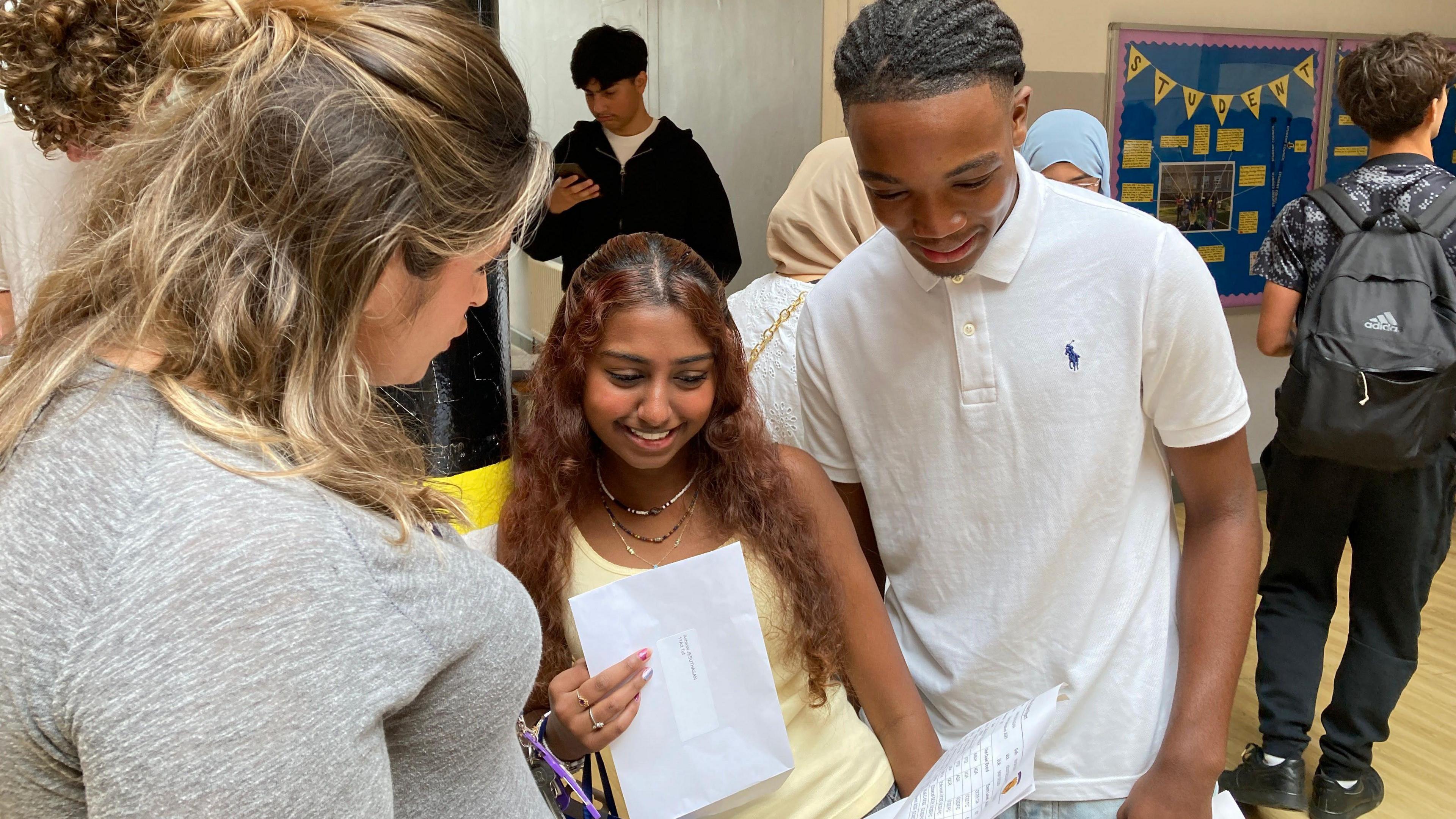 Lamari and Ashwini are standing next to each other, looking at a piece of paper with a woman who looks like their teacher. Lamari wears a white Polo Ralph Lauren t shirt and jeans. Ashwini wears a pale yellow tank top with a stack of different chain necklaces, she has long red wavy hair. Their teacher is a woman with mid length blonde hair, wearing a grey marl long sleeved t shirt. They are standing in a school hall with other students in the background.