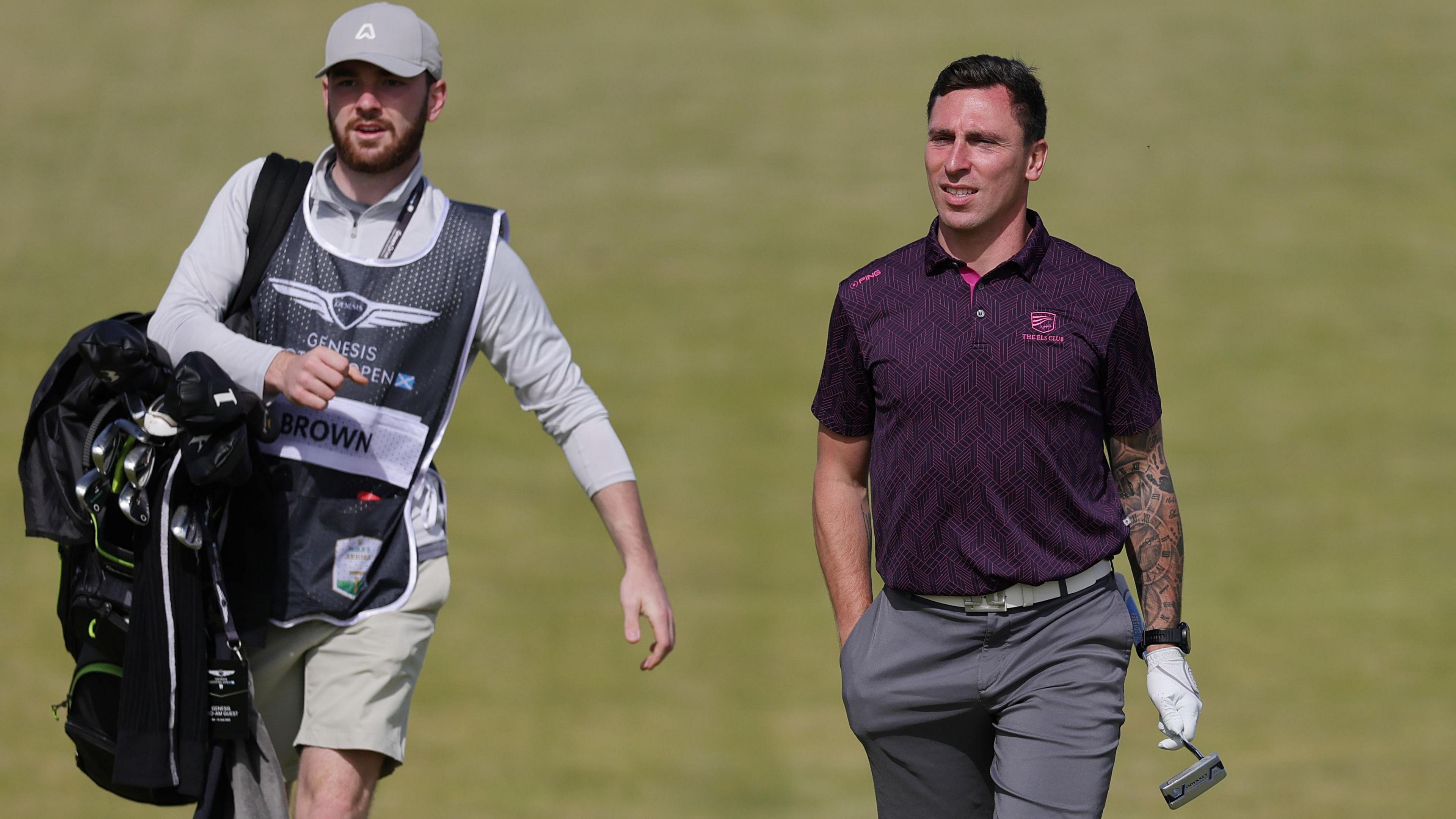 Scott Brown, who is wearing a maroon polo shirt and dark grey shorts, walks along the fairway carrying his putter. A caddy, wearing a long sleeved grey top is walking to his right carrying a black golf bag. He is also wearing a silver cap.