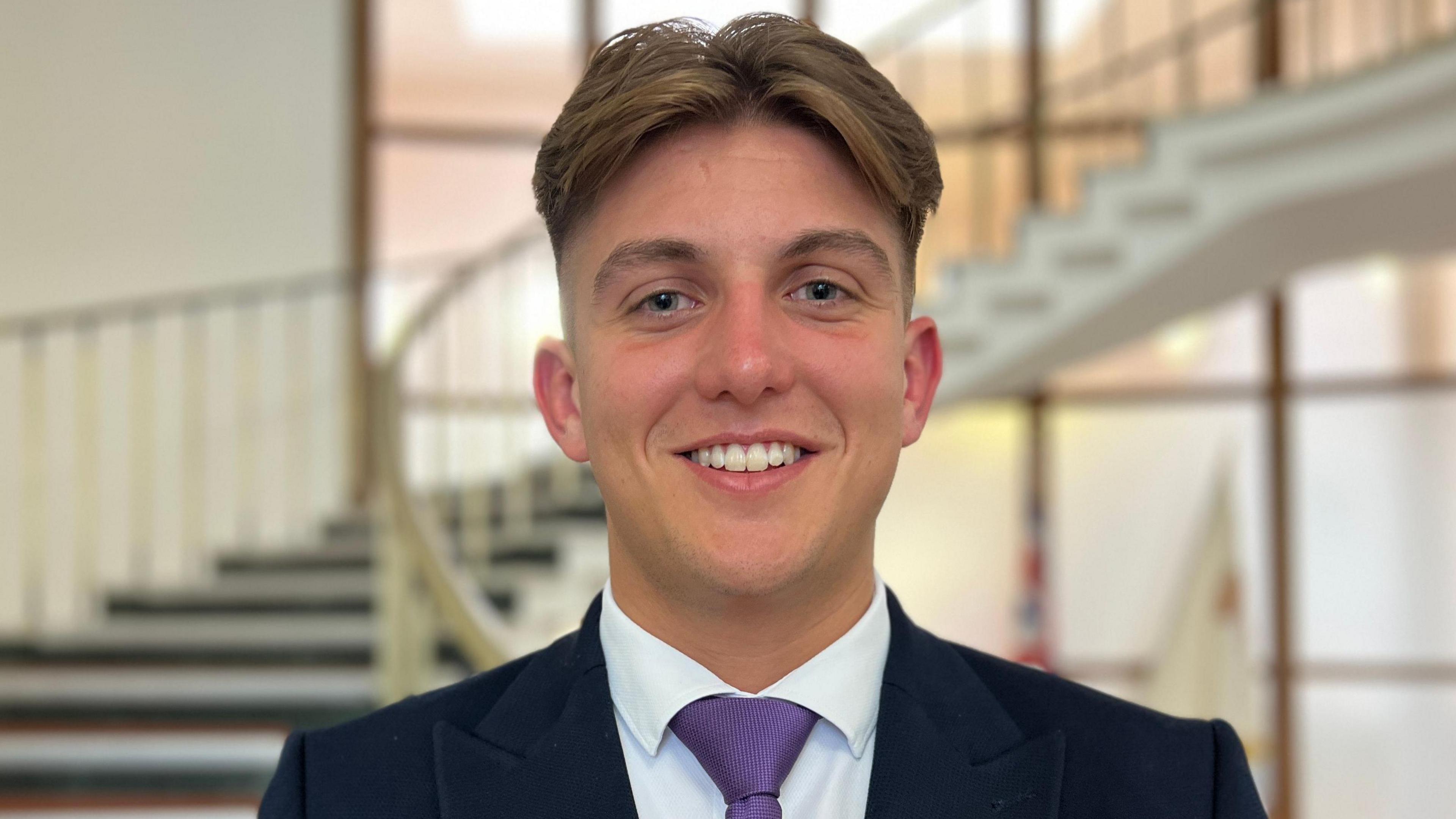 A man with brown hair, cropped at the sides, smiles at the camera in front of a curved staircase in a council building. He is wearing a black suit, white shirt and purple tie. 
