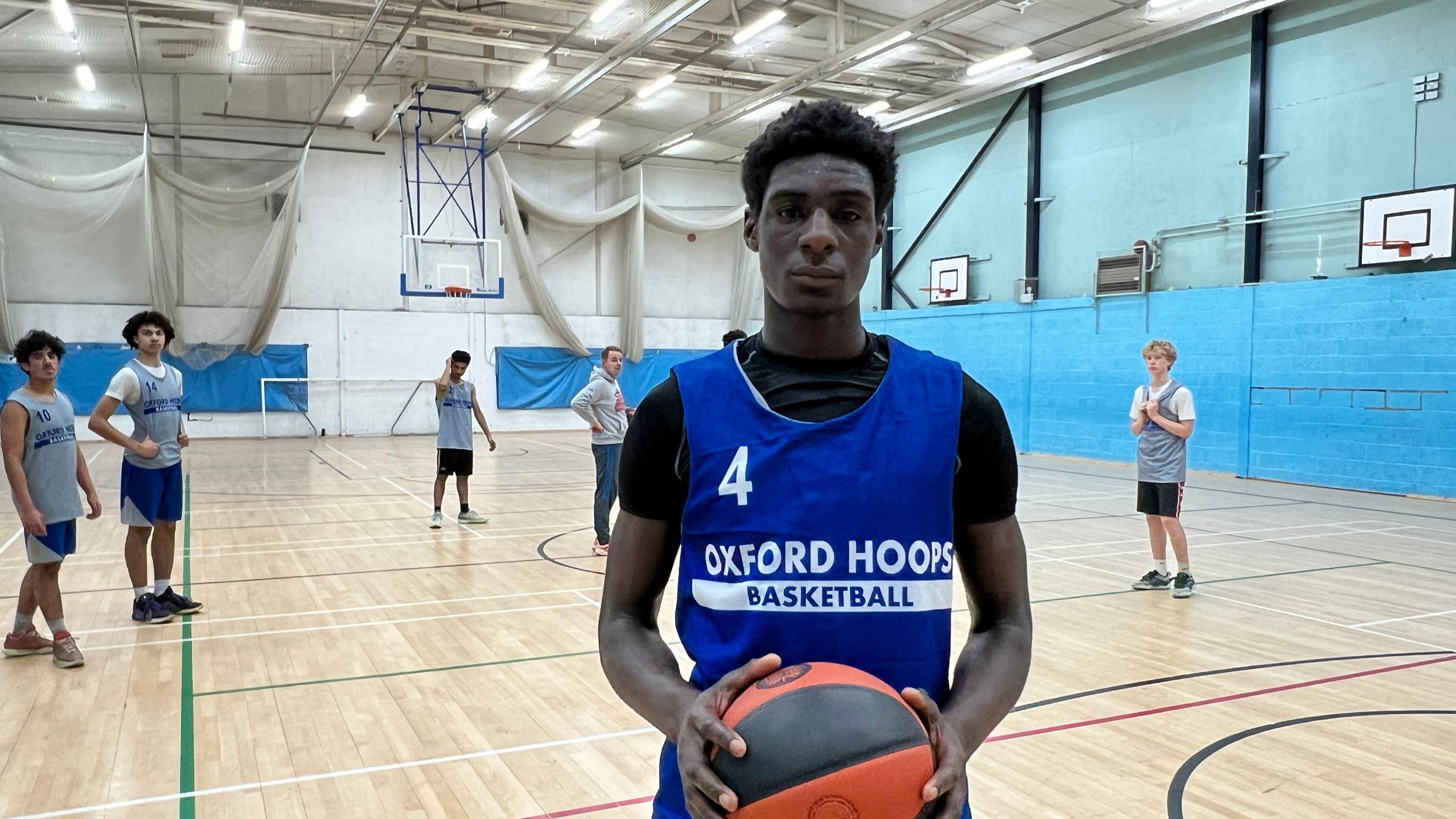 David Okerike faces the camera. He's wearing a blue vest with 'Oxford Hoops' printed on it in white letters. He's holding an orange and black basketball in both hands. Behind him is a school hall, with a wooden panelled floor marked for various sports and several basket ball hoops fixed to the walls. Behind David there are several other young players dotted around the court looking in the direction of the camera.