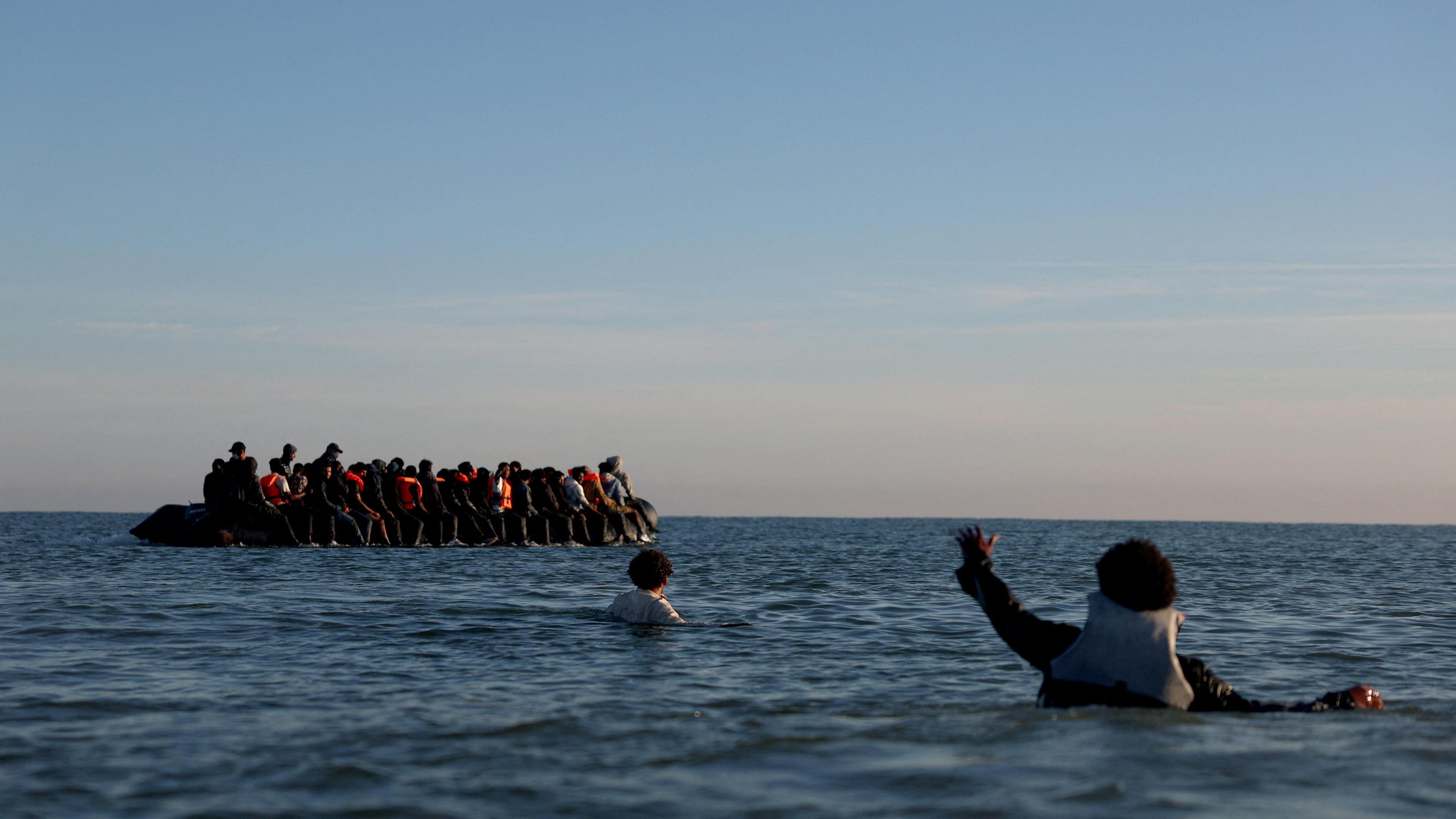 A group of migrants on an inflatable dinghy leave the coast of northern France in an attempt to cross the English Channel to reach Britain, from a beach in northern France. In the foreground of the picture there are two people who are in the water. One is holding out their hand to towards the dinghy