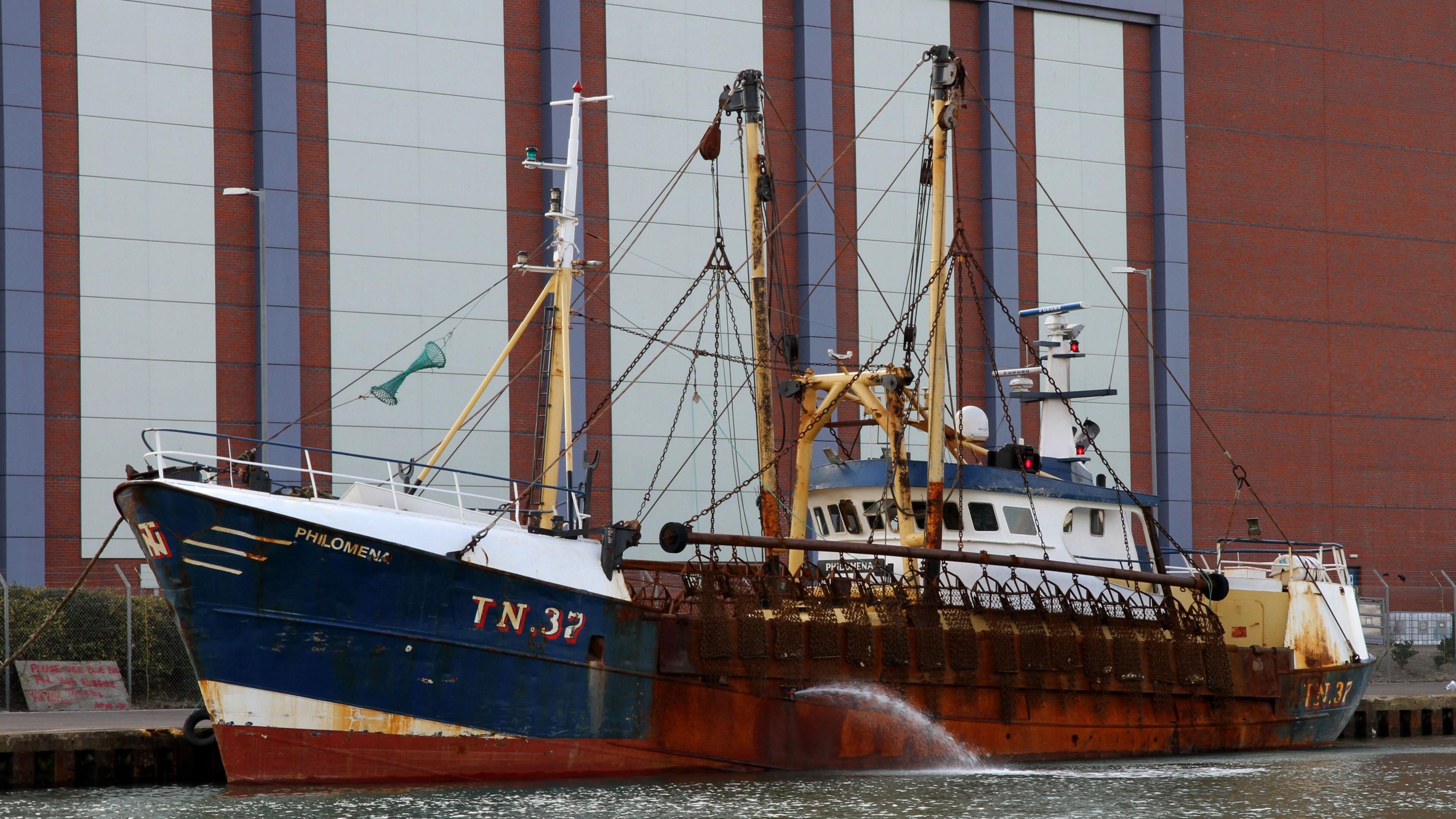 A blue and white fishing boat called the Philomena. It is very rusty. It is in a dock with a large modern building behind.