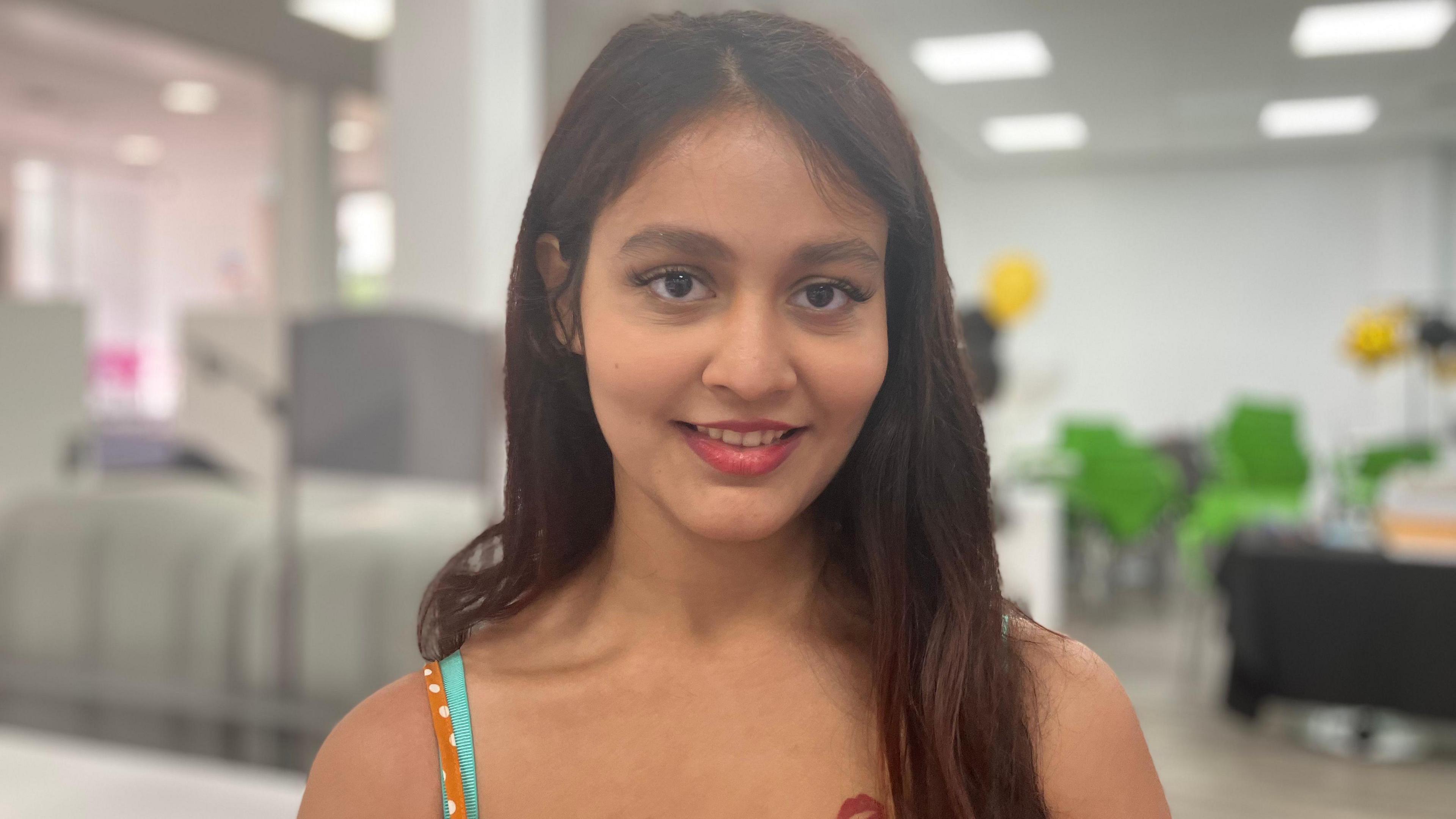 Dipa looks at the camera, smiling. She has long brown hair and wears red lipstick. She has a strappy camisole top on, which is orange with white polka dots. The background behind her is blurred, but shows an area of the college with some green desk chairs.