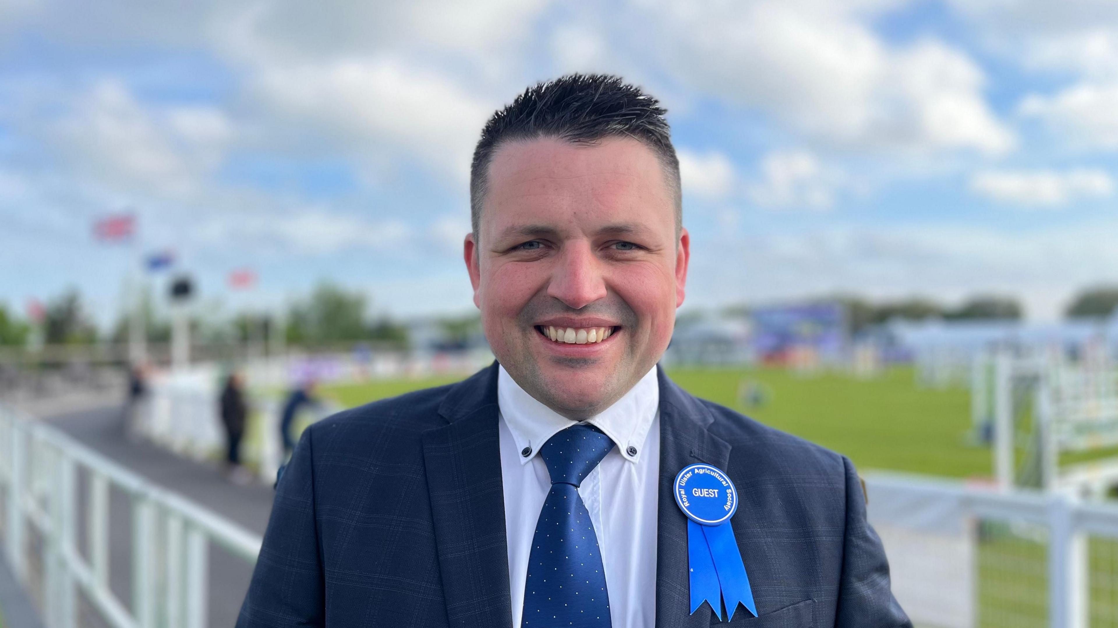 Richard Beattie. He has shirt dark hair, wearing a navy blazer, white shirt, blue tie and blue rosette pin.