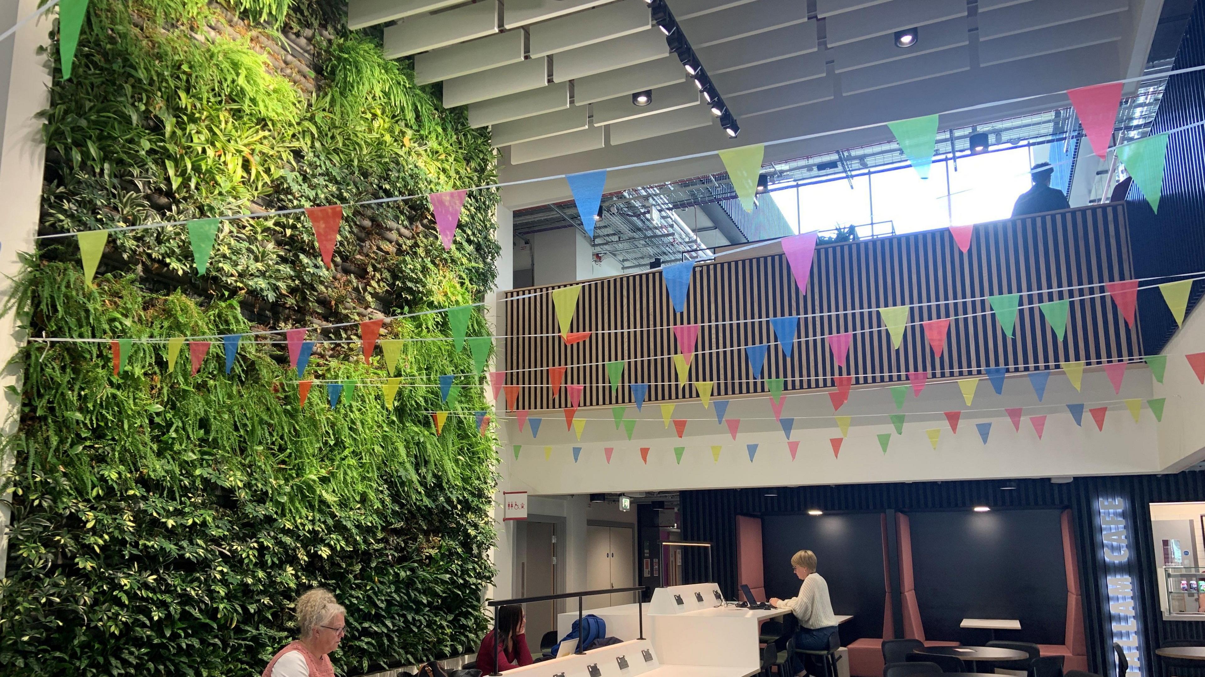 A wall is covered floor to ceiling in real living green plants and foliage. Is overlooks a bank of tables and there is multi-coloured bunting across the ceiling