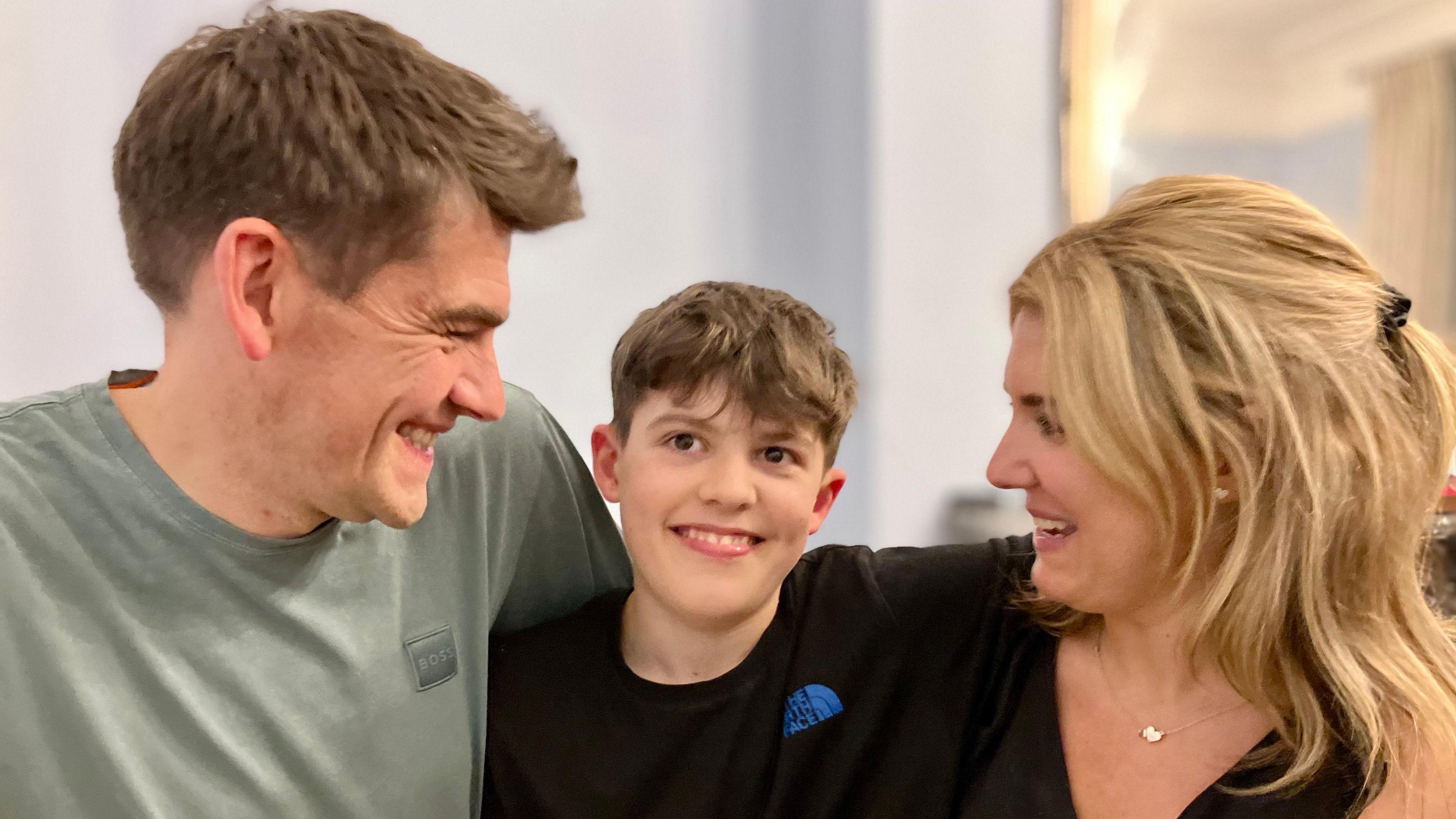 Dan Burrows wearing a grey t-shirt with his arm around his son Lucas, wearing a black t-shirt, and his wife Lucy wearing a black top. Both parents looking at their son and smiling