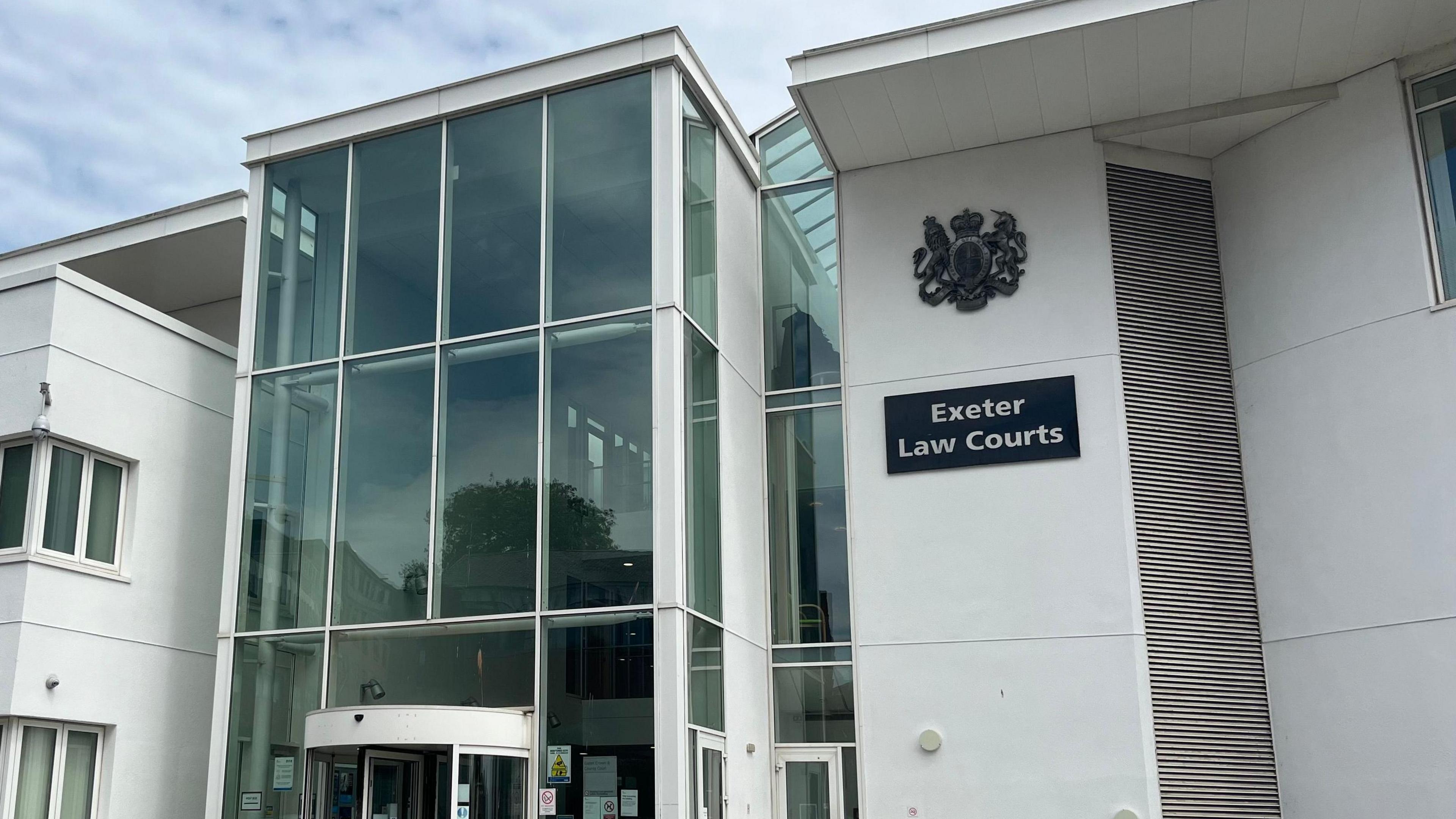 The exterior of Exeter Crown Court. The front of the building is white and has large glass windows on the left. On the right, there is a black sign with the words 'Exeter Law Courts' with a badge above.