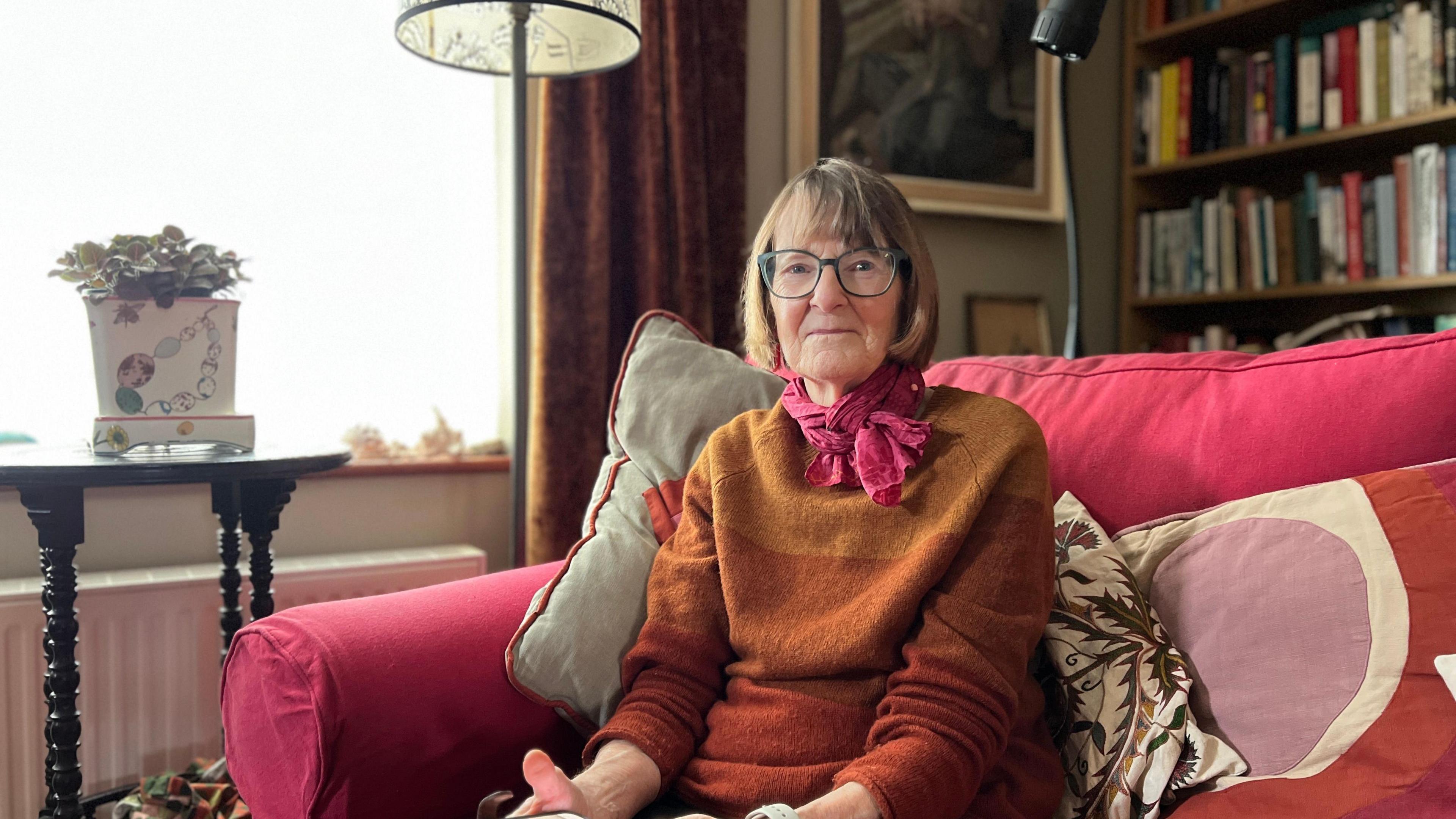 Sarah Carter is wearing an orange scarf over a jumper. She is sitting on a pink arm chair with book shelves behind her