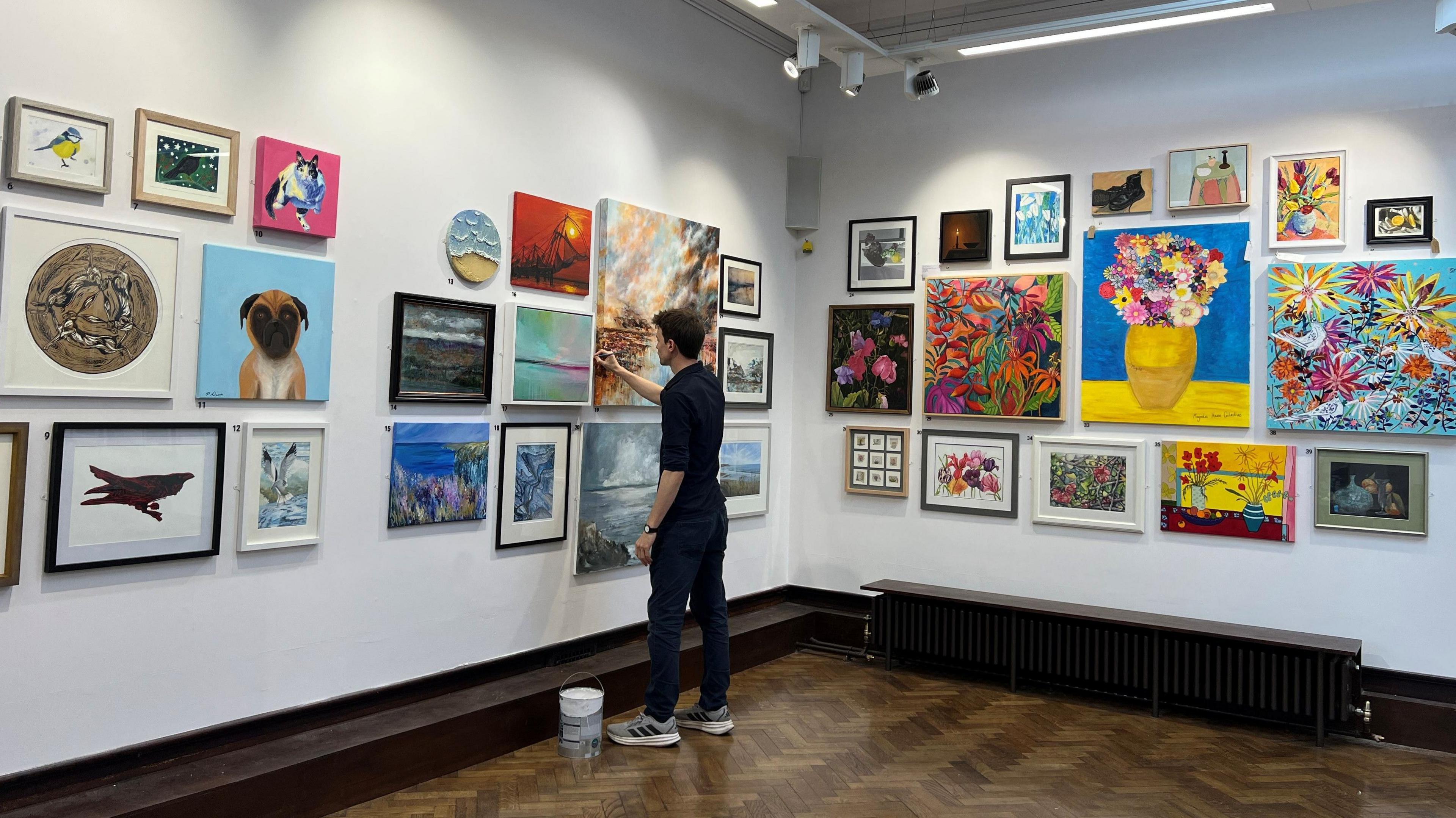 A man standing in a gallery space with an array of colourful artwork on the white walls. Among them is a picture of some brightly coloured flowers in a vase, as well as an image of a brown and white dog.