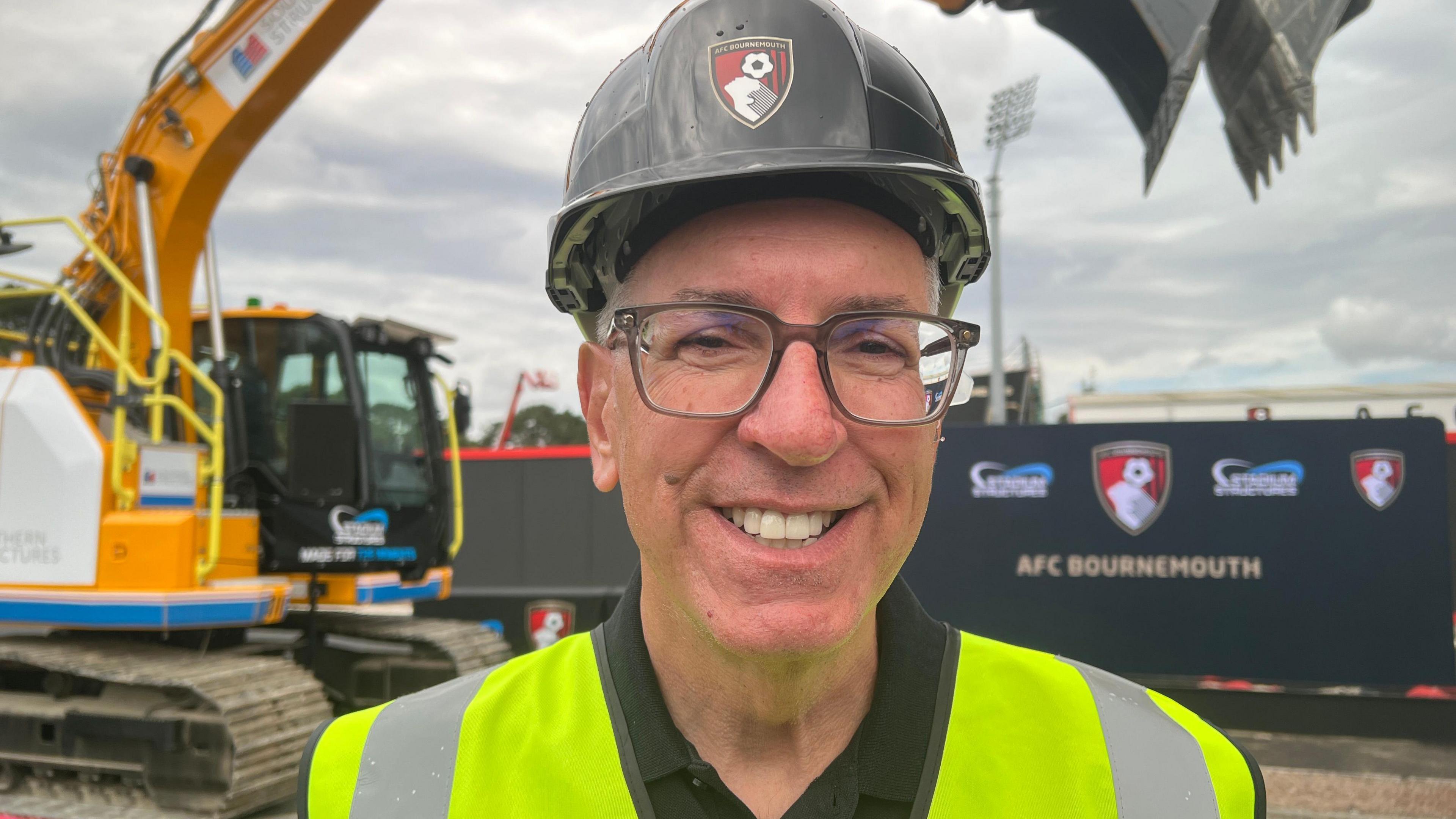 Jim Frevola wearing a high visibility vest and hard hat with AFC Bournemouth branding. In the background the stadium can be seen along with an excavator. 