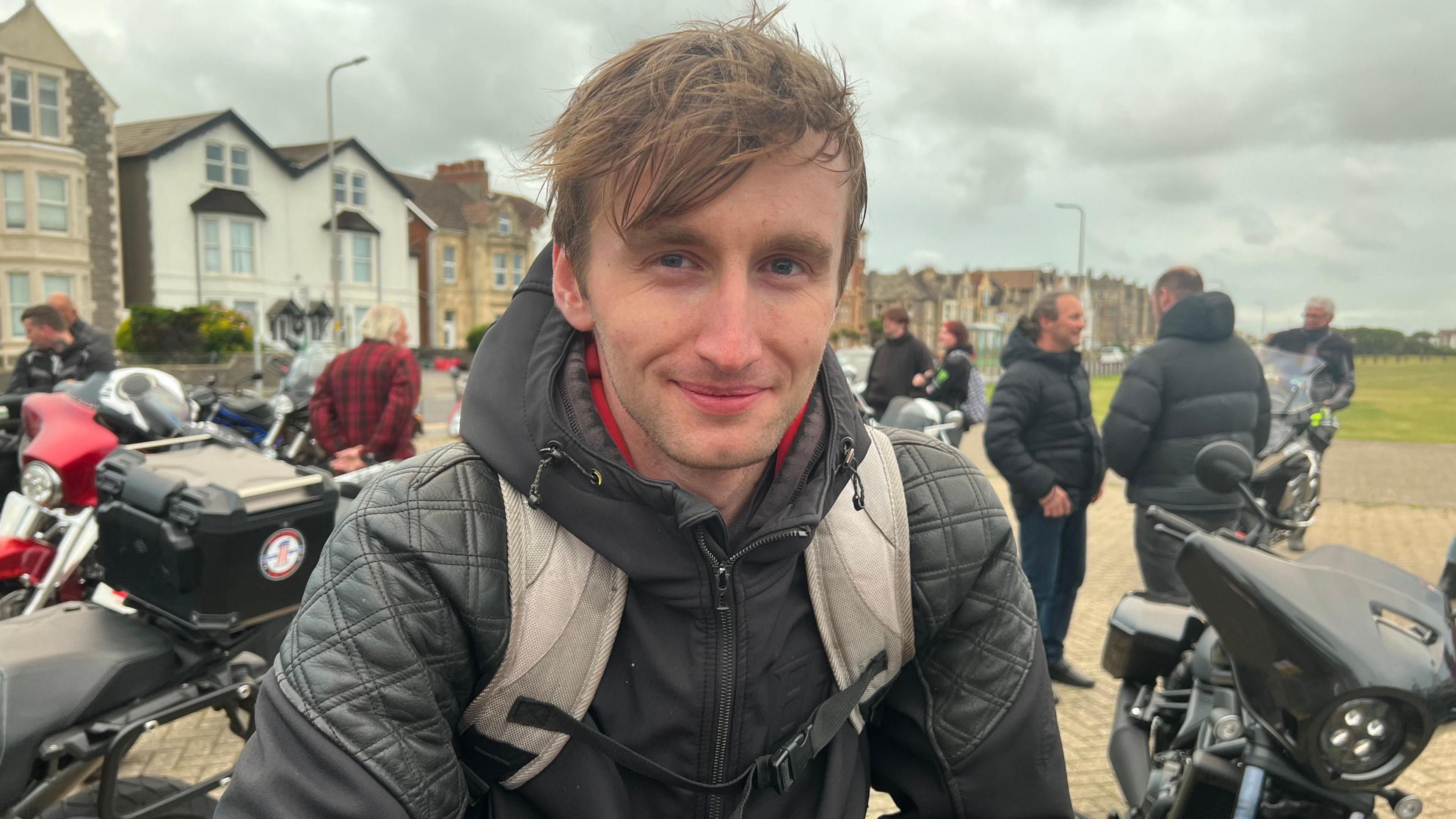 Mykel wearing a dark hoodie  and a grey backpack with short light brown hair smiling at the camera. There are people loitering in the background in a car park with a few other motorbikes in view.